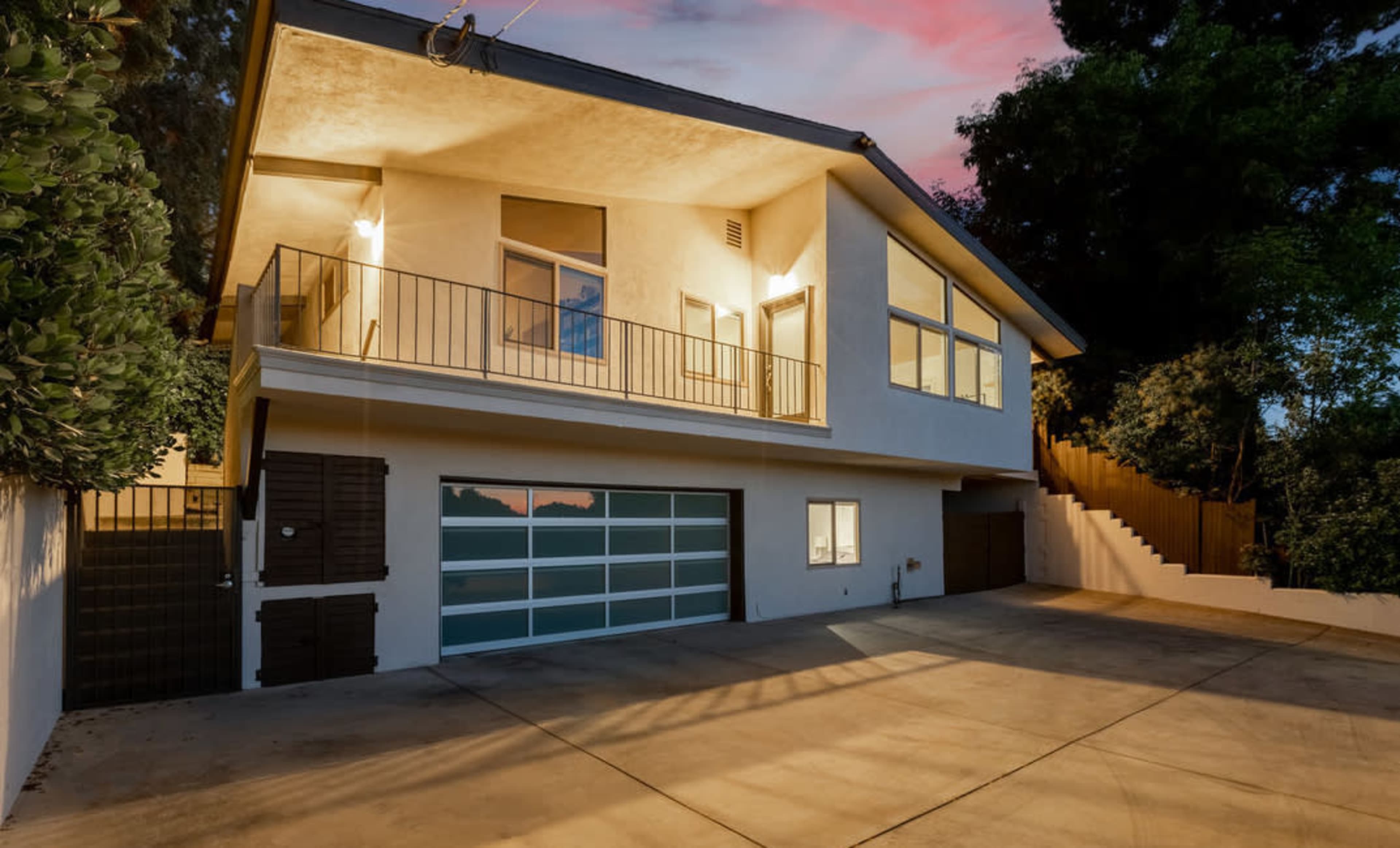 A two-story house with a balcony and a garage is situated on a concrete driveway surrounded by greenery.