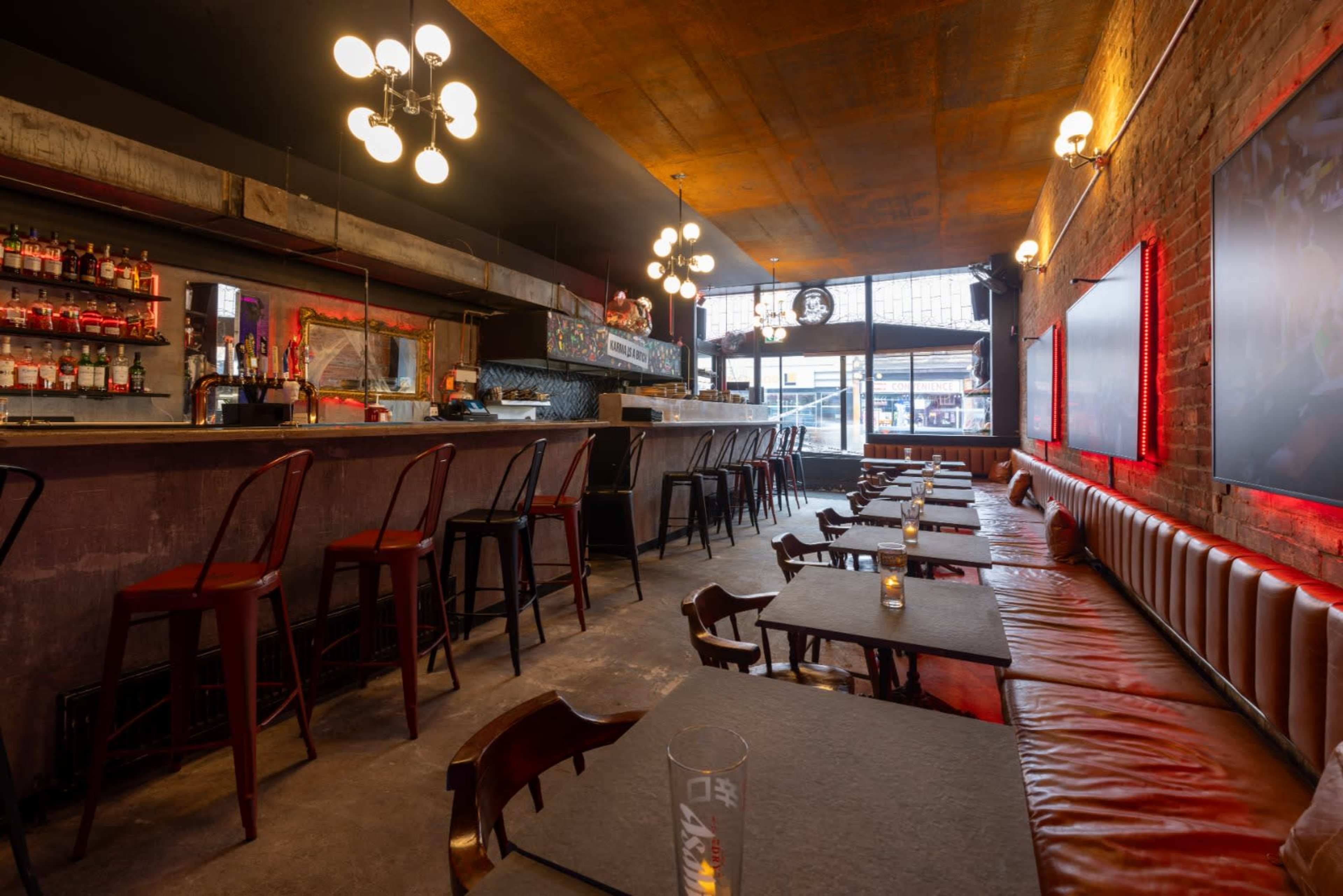 The image shows a dimly lit bar with a long countertop, high stools, and rows of tables along a leather banquette.