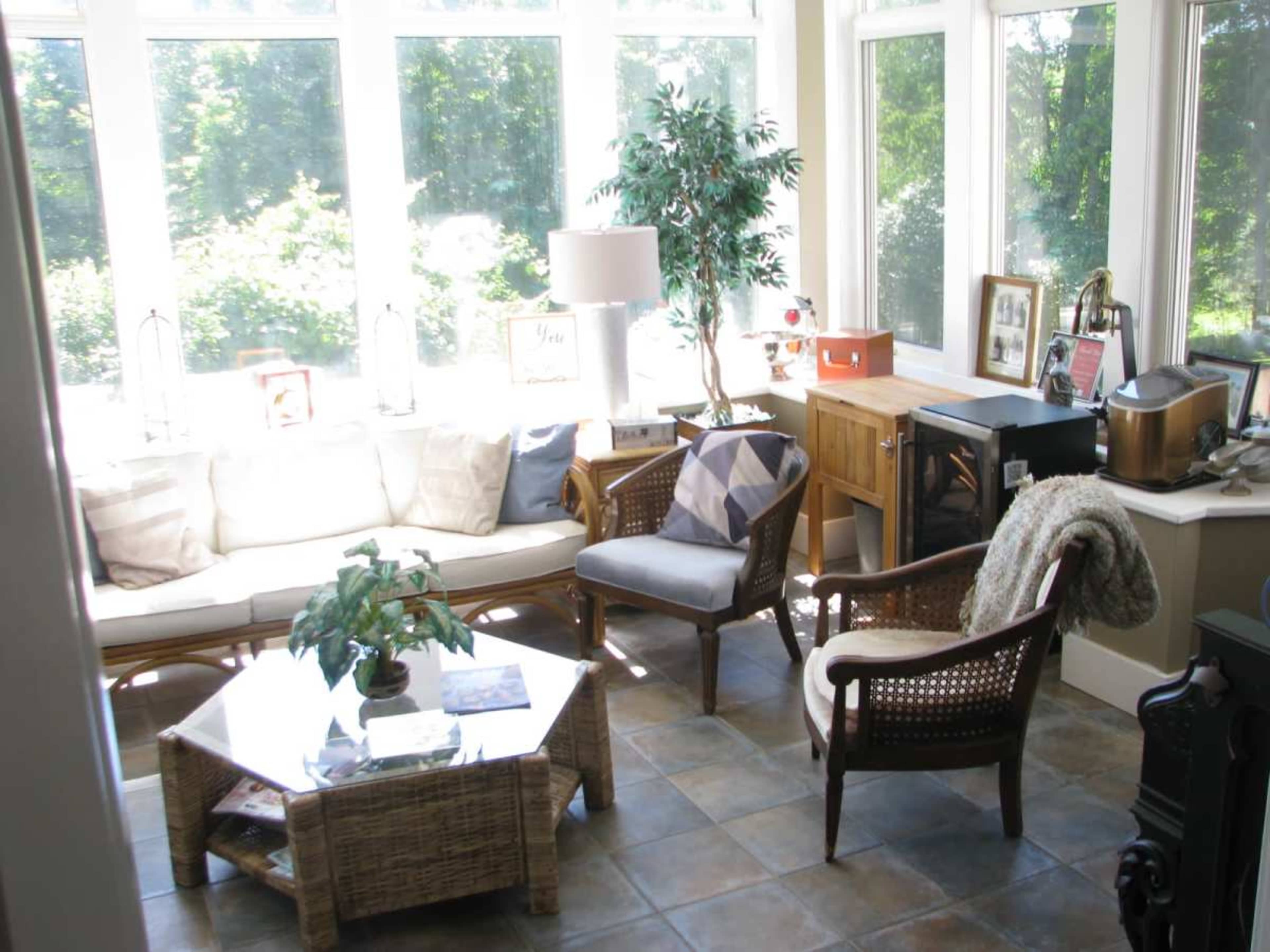 A sunlit room with large windows, featuring a seating area with a sofa, a chair, and a coffee table, along with a small wooden cabinet and greenery.