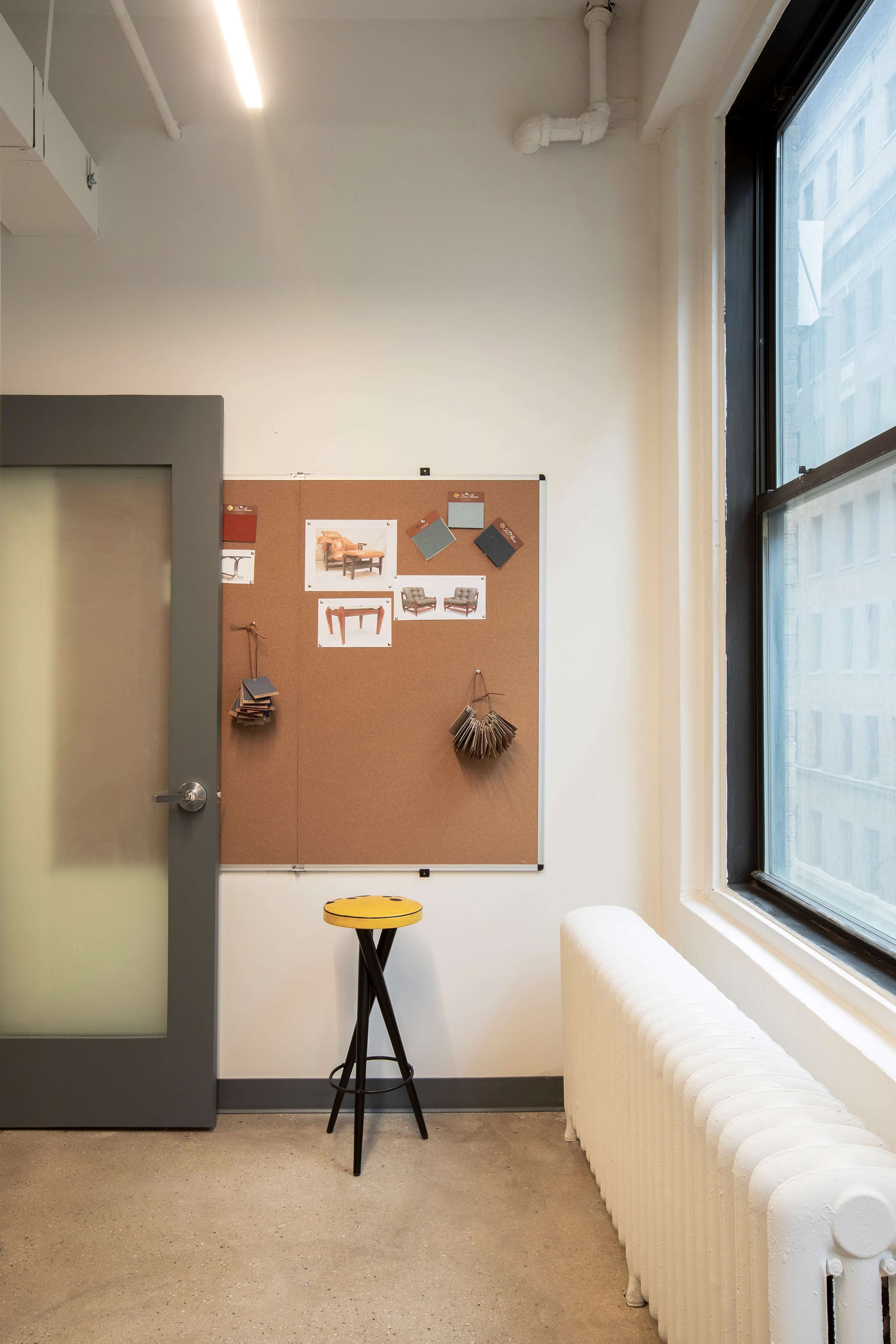 A corkboard with various images and materials is mounted on a wall next to a window, with a small yellow stool placed nearby.