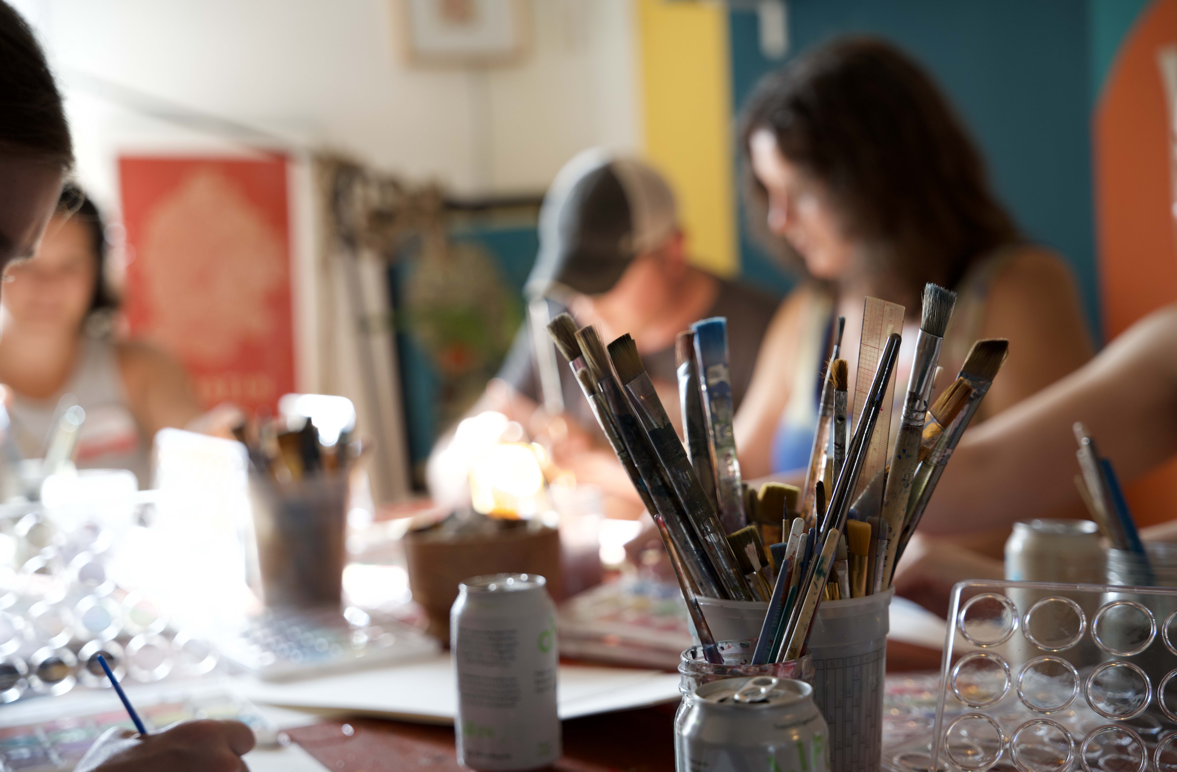 A group of people is engaged in a painting activity at a table filled with art supplies and drinks.
