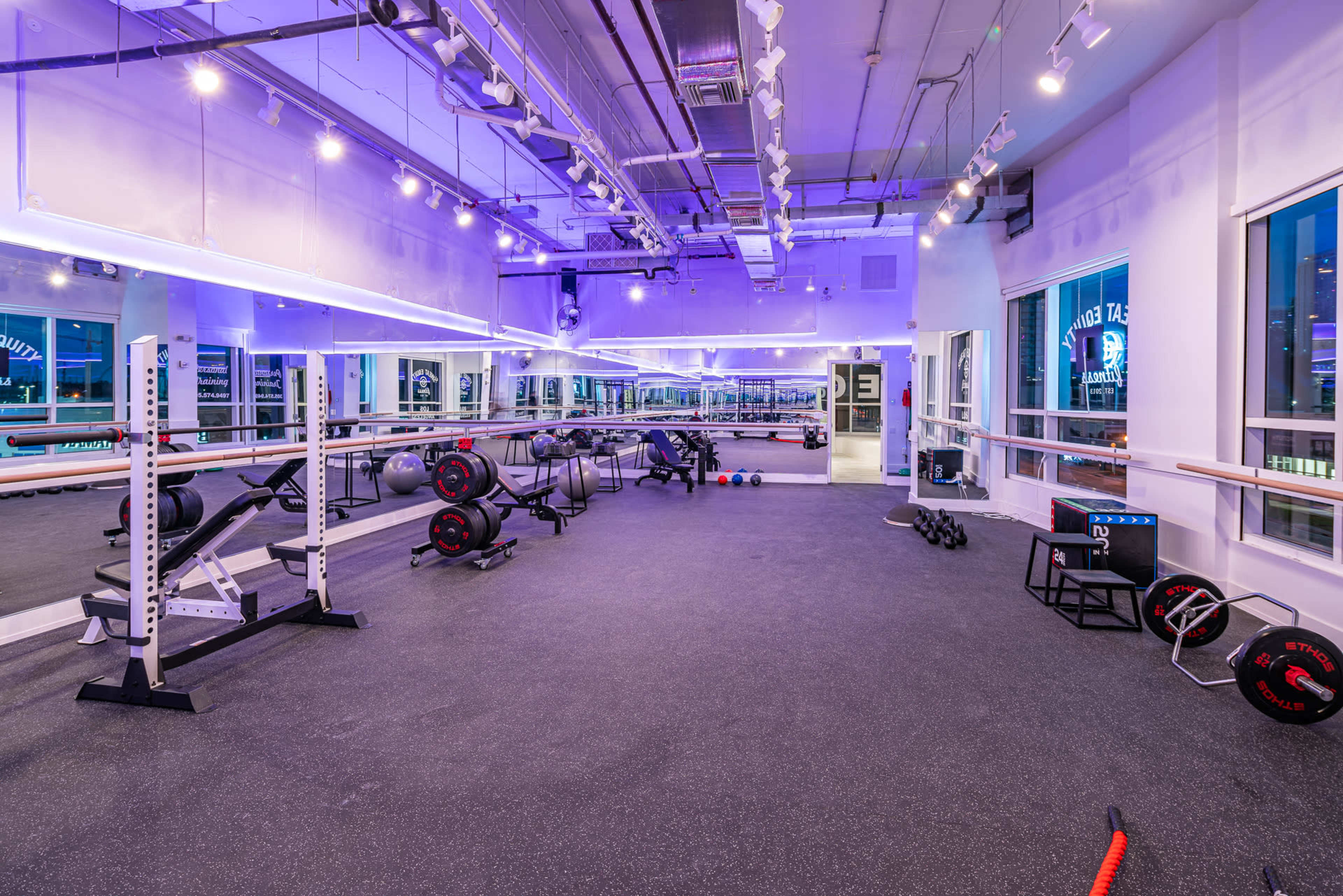 A spacious, modern gym with various weightlifting equipment, exercise mats, and mirrors under purple lighting.