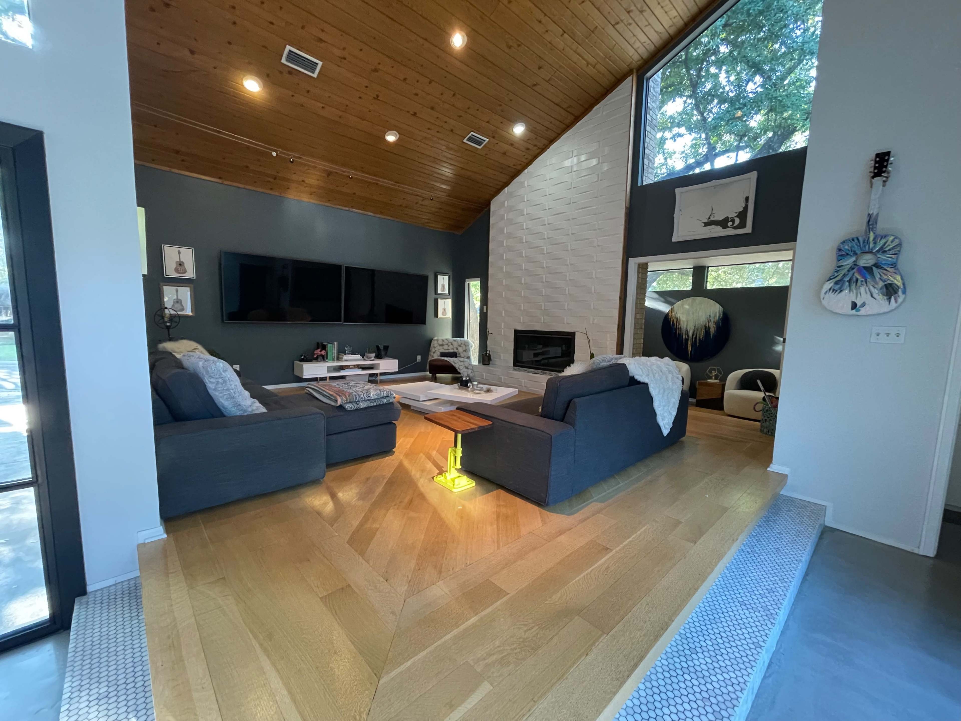 A modern living room with two gray sofas facing a coffee table, large windows, and a fireplace, featuring a mix of wooden and tiled flooring.