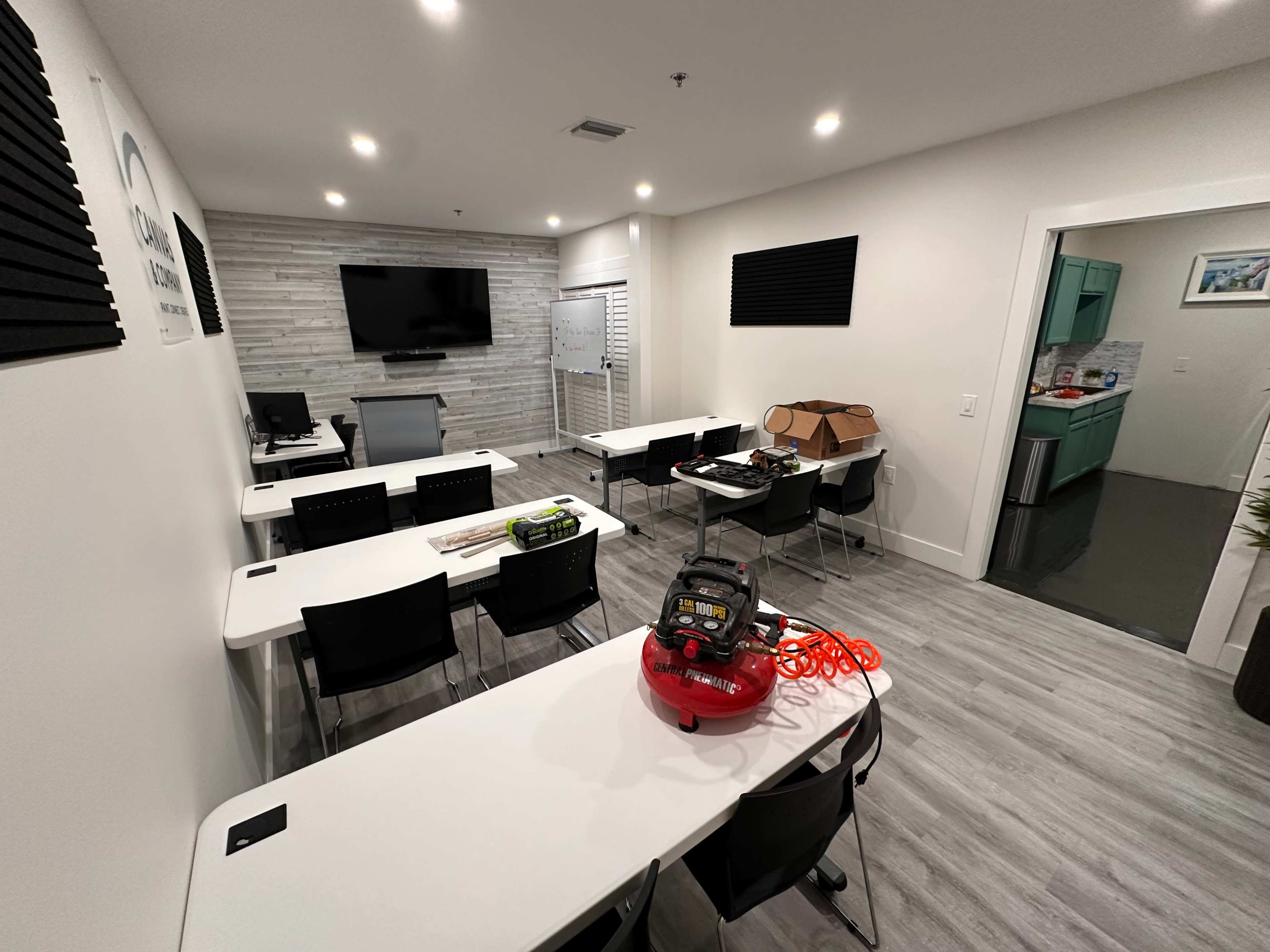 The image shows a modern training room set up with several white tables and black chairs arranged in a U-shape, featuring a large television screen and a kitchenette in the background.