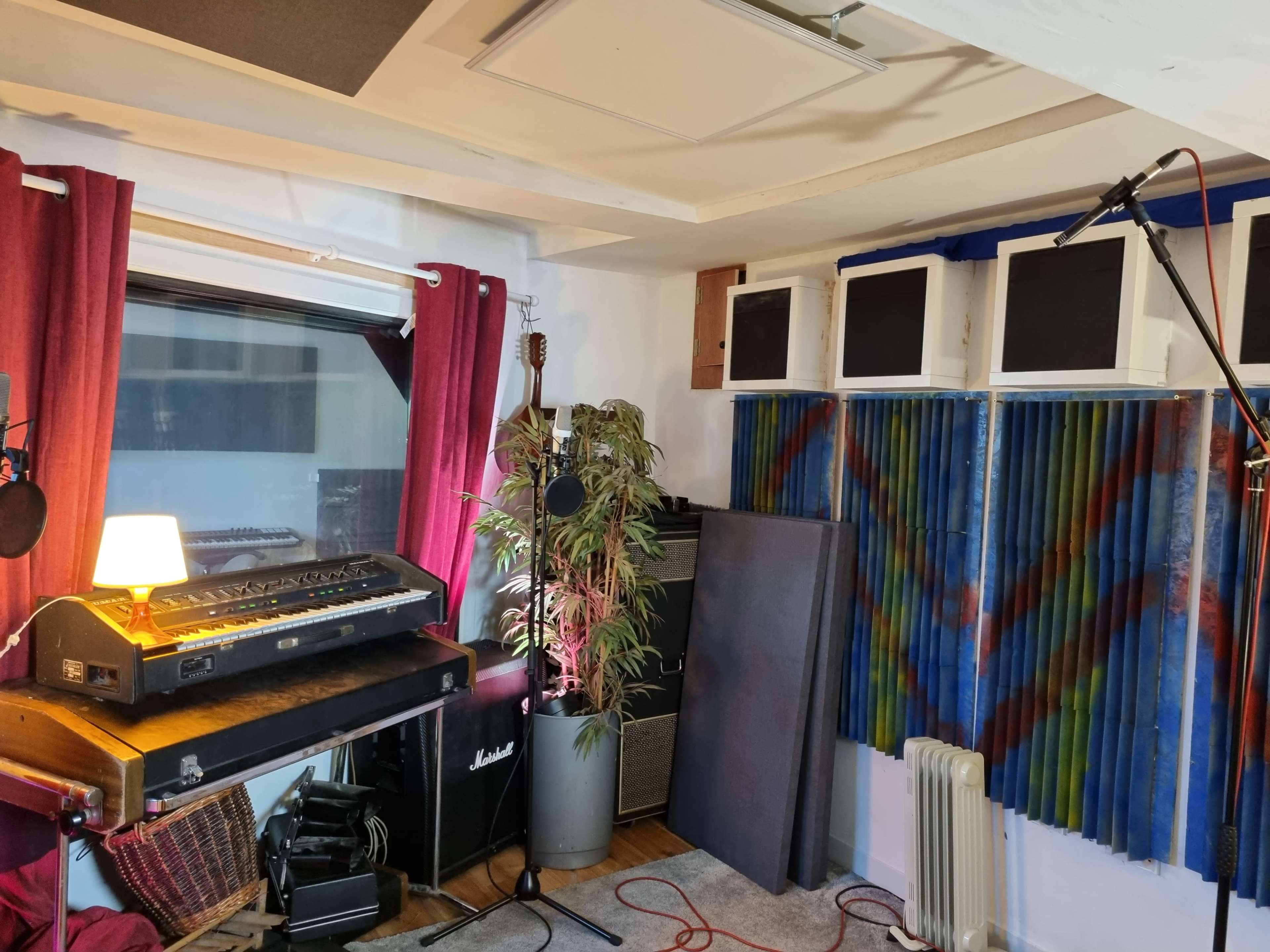The image shows a music recording studio featuring a vintage keyboard, acoustic panels, a microphone, and various equipment arranged in a brightly lit space with red curtains.
