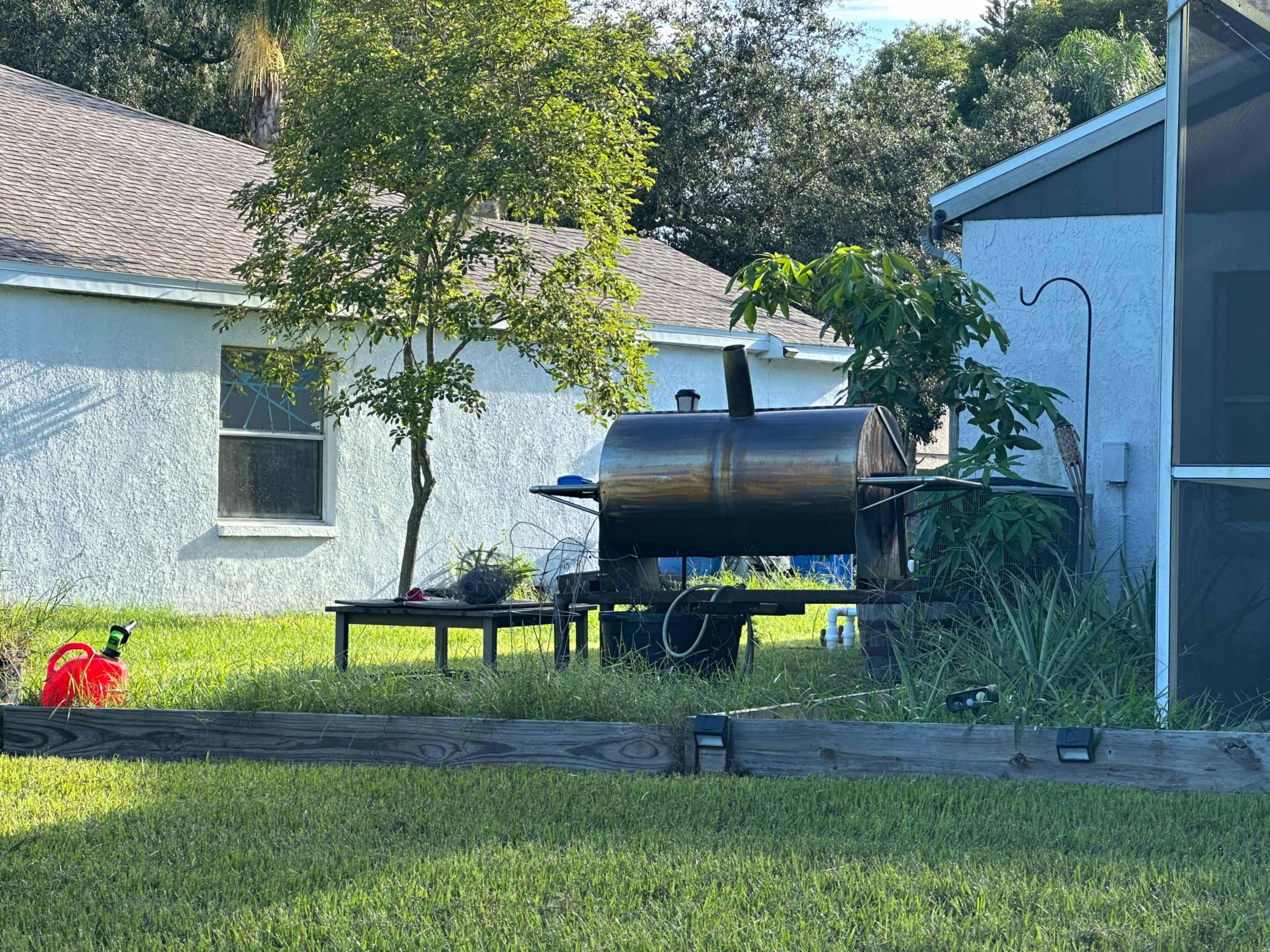 A black smoker grill sits on a table in a grassy backyard, surrounded by a tree and a few plants.