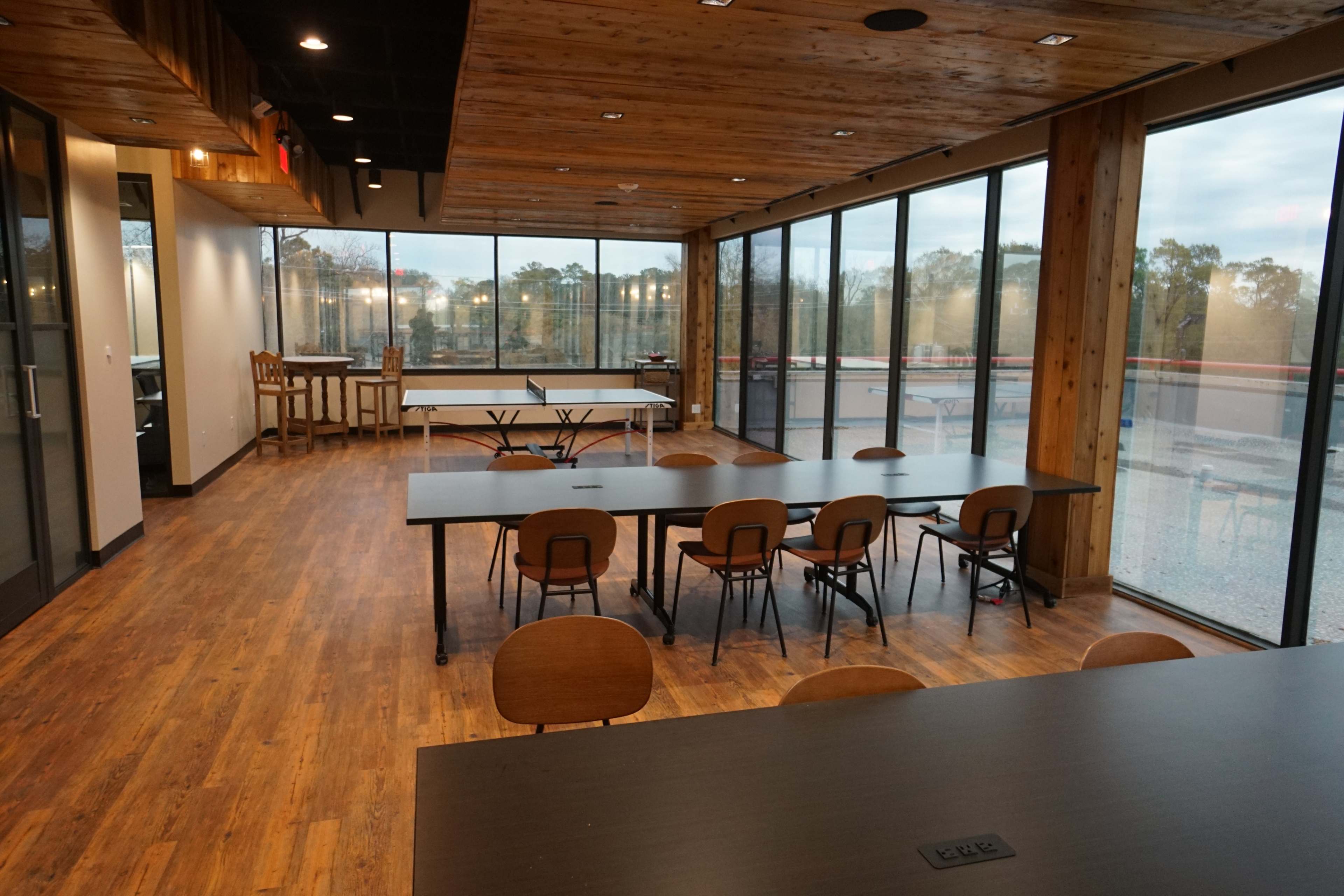 The image shows a spacious room with wooden flooring, large windows, multiple tables, and chairs arranged for work or meetings.