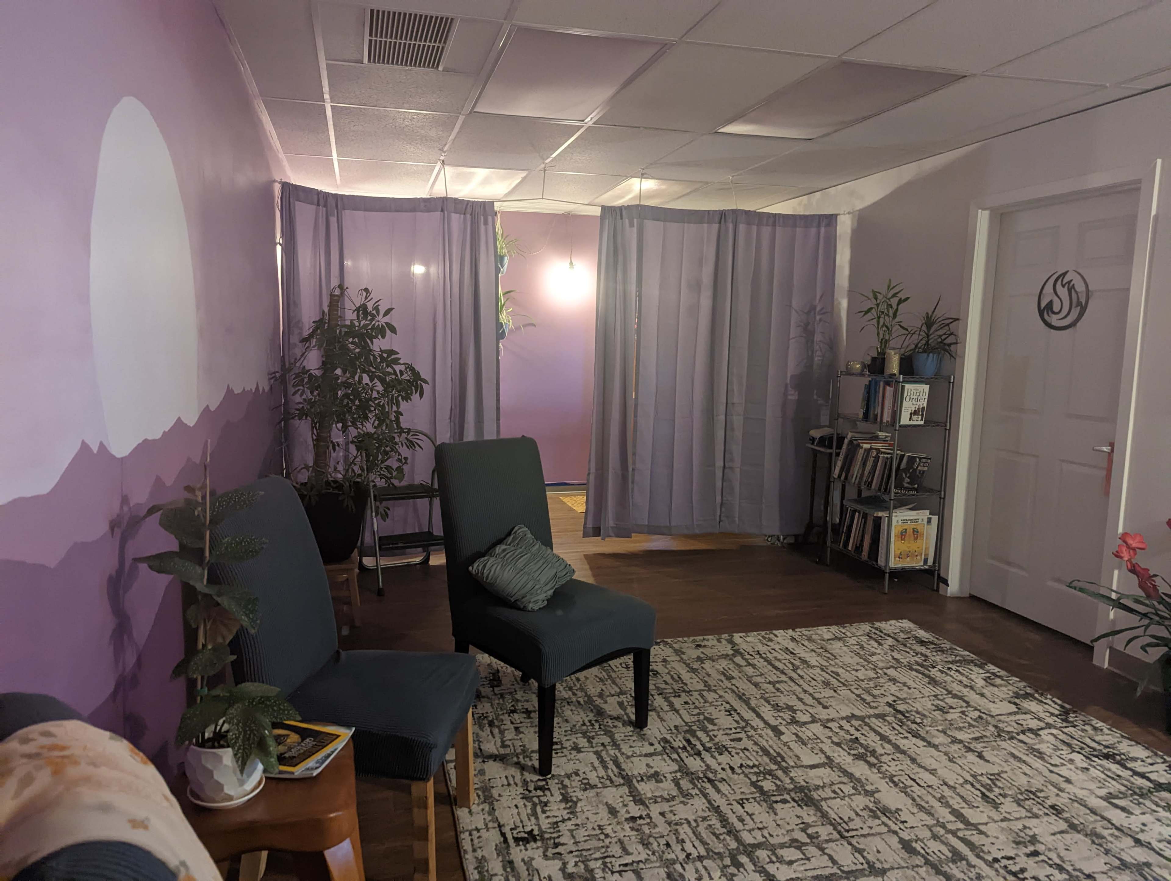 A waiting area with two chairs, a bookshelf, and plants, all set against a purple wall with curtains separating different sections.