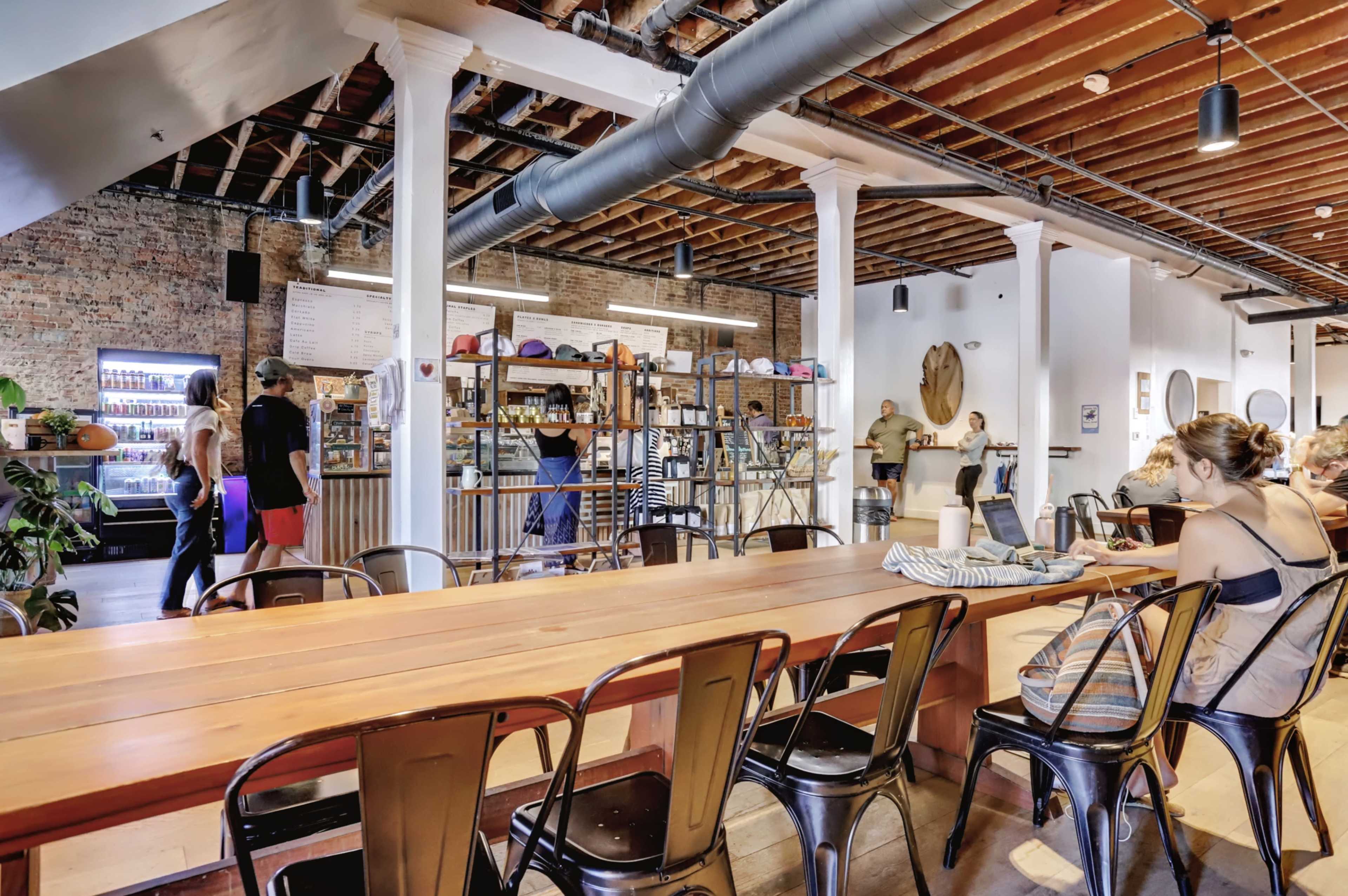 A modern café features a spacious interior with wooden tables, exposed brick walls, and patrons engaging at various seating areas.