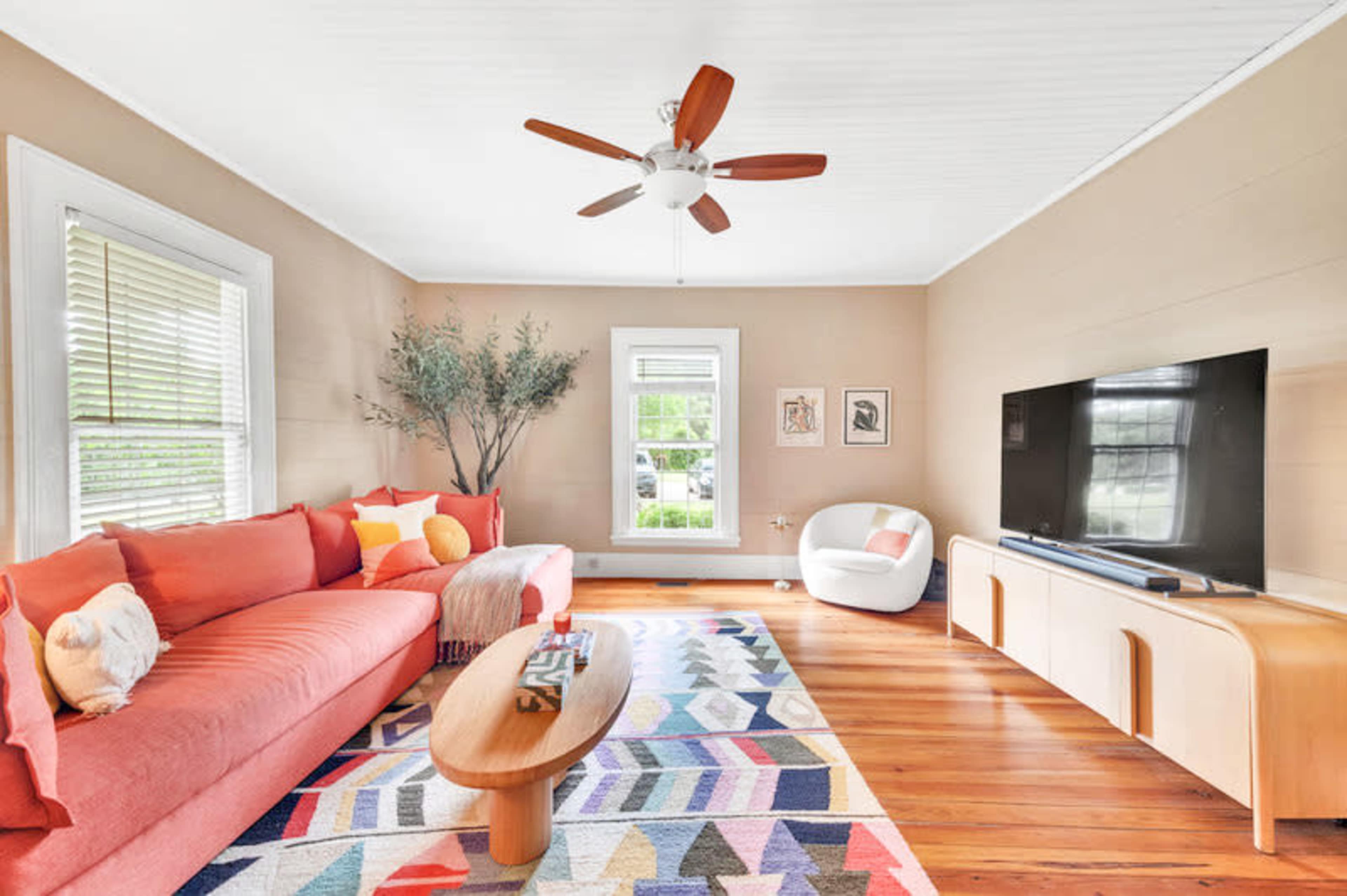 The living room features a large coral sofa, a round wooden coffee table, a colorful patterned rug, and a television on a light wood cabinet, with a window allowing natural light to enter the space.