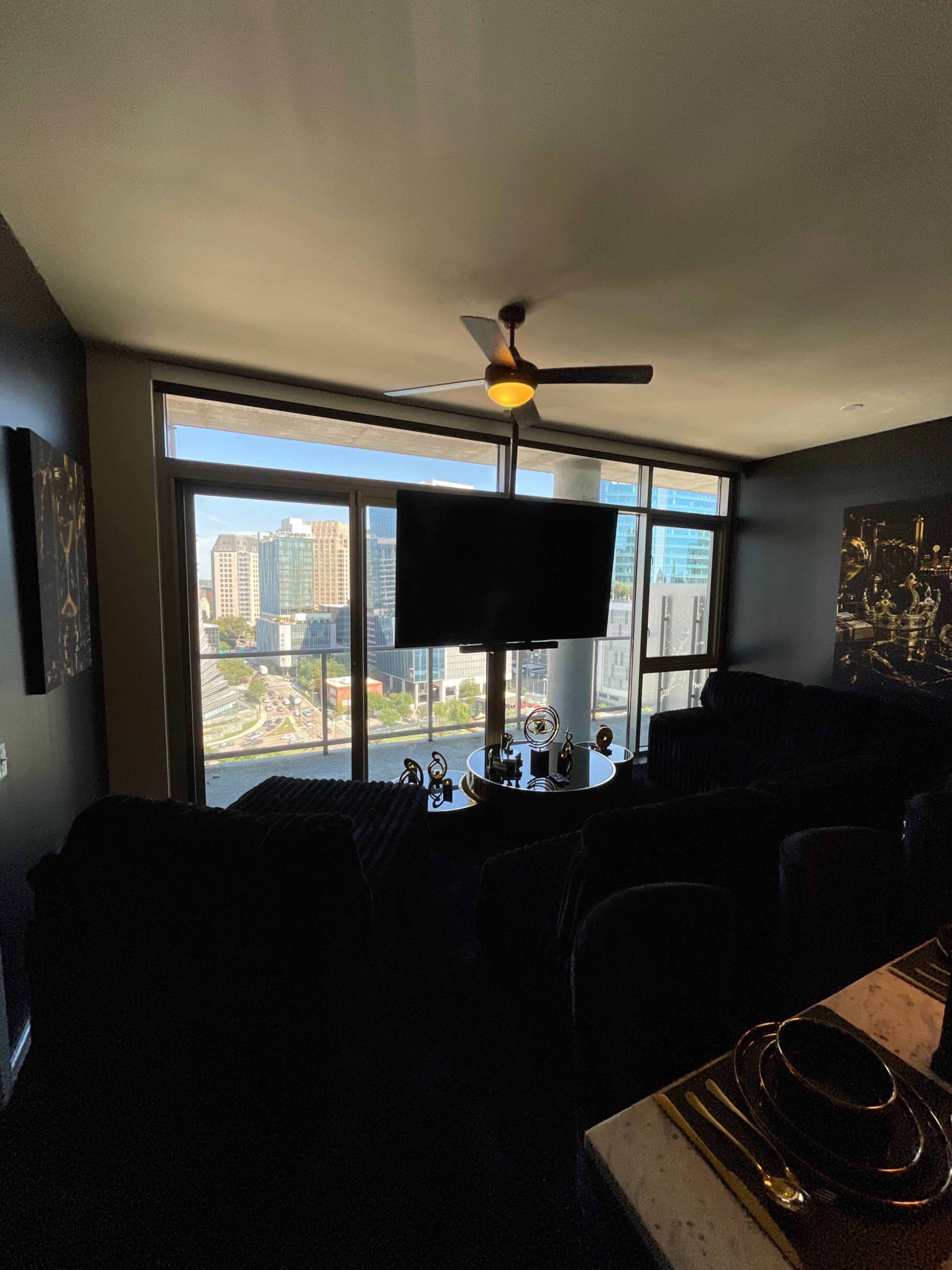The image shows a modern living area with a large window overlooking city buildings and a black couch arrangement facing a mounted television.