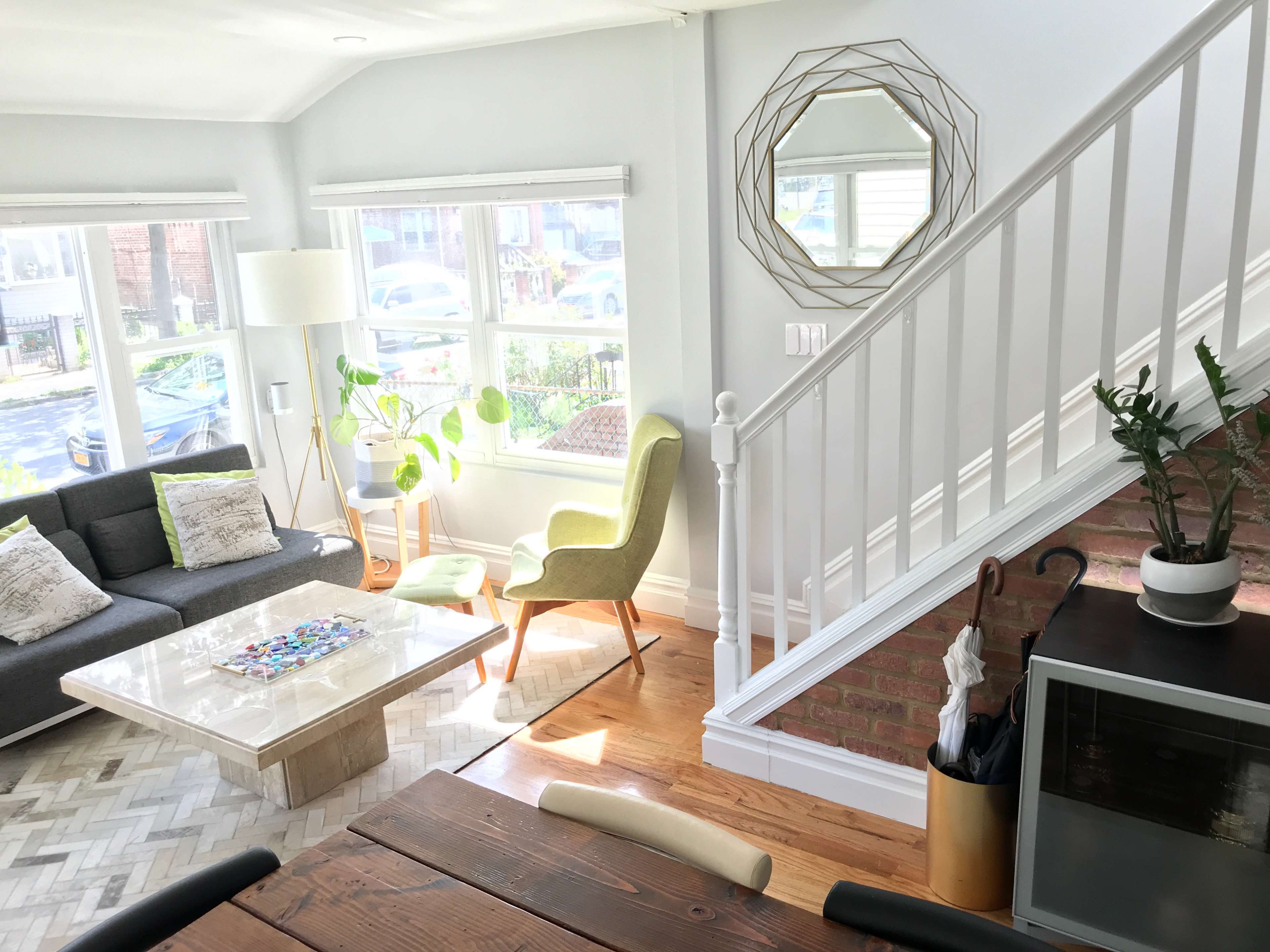 The image shows a living room with a gray sofa, a coffee table, a green chair, and a staircase in a well-lit space with large windows.