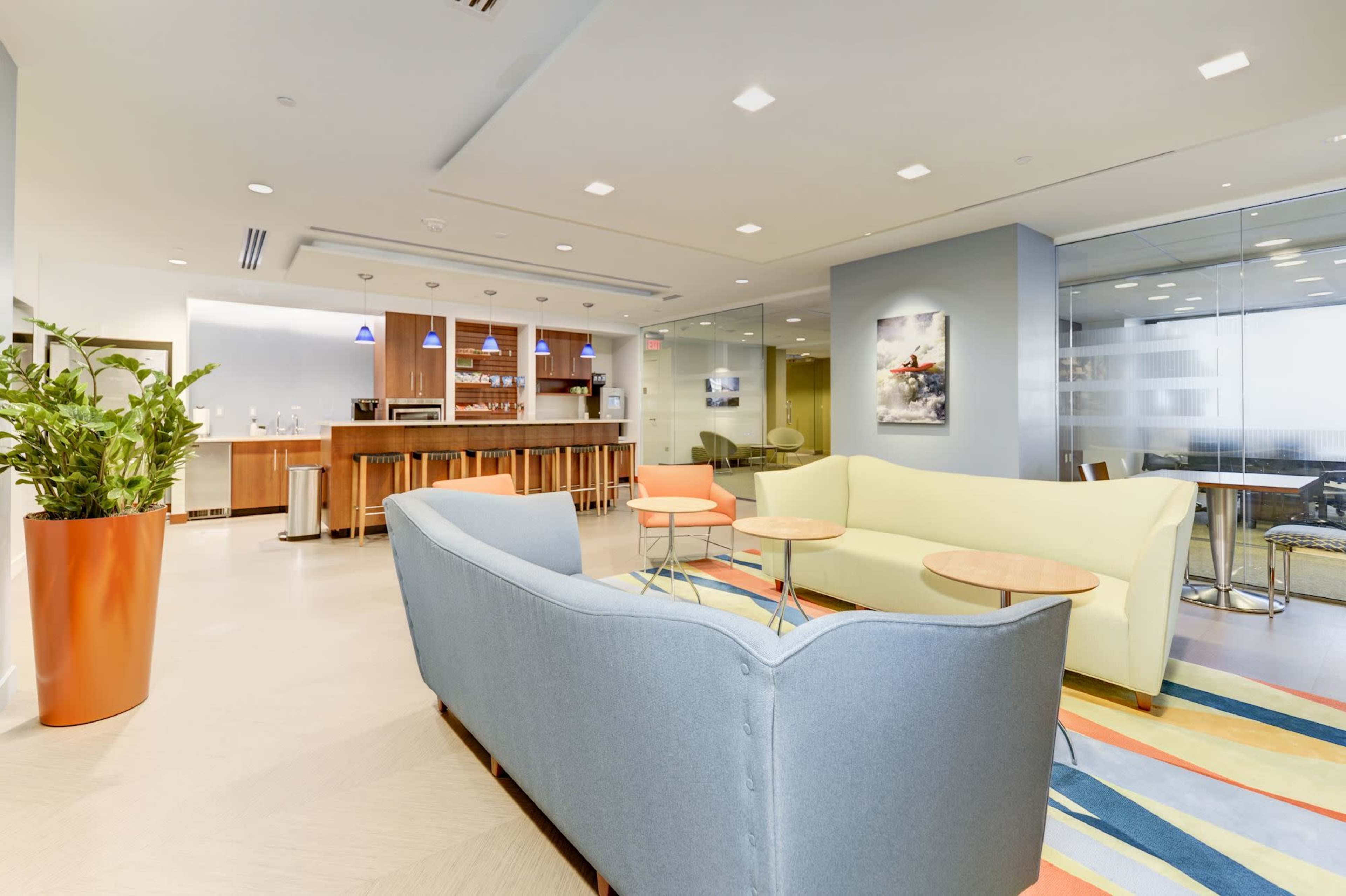 The image shows a modern office lounge area with a light blue sofa, orange and beige accent chairs, and a kitchenette in the background.