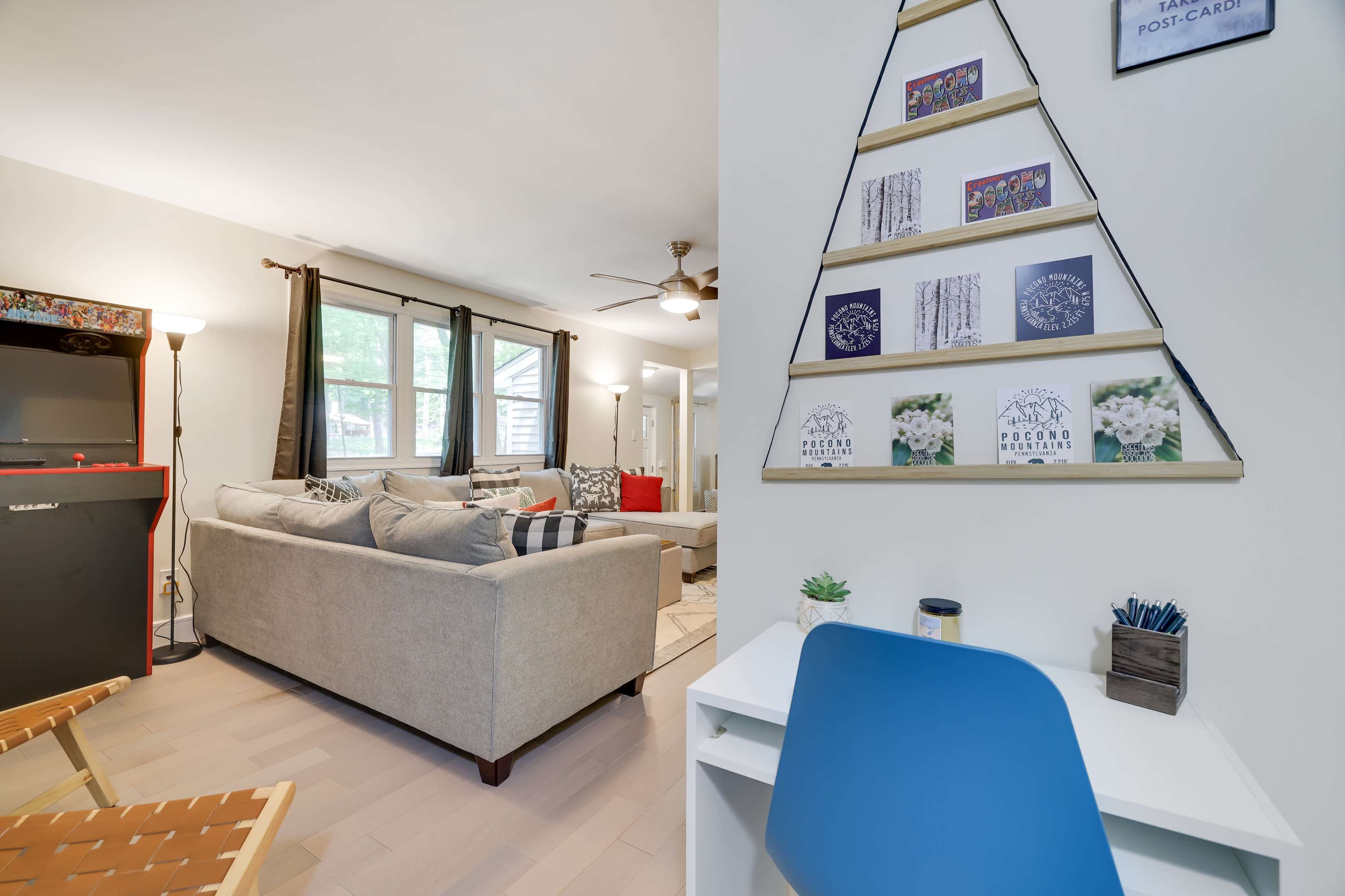 A modern living space features a light-colored sofa and armchair, framed by large windows, with a decorative wall displaying cards and a small desk in the foreground.