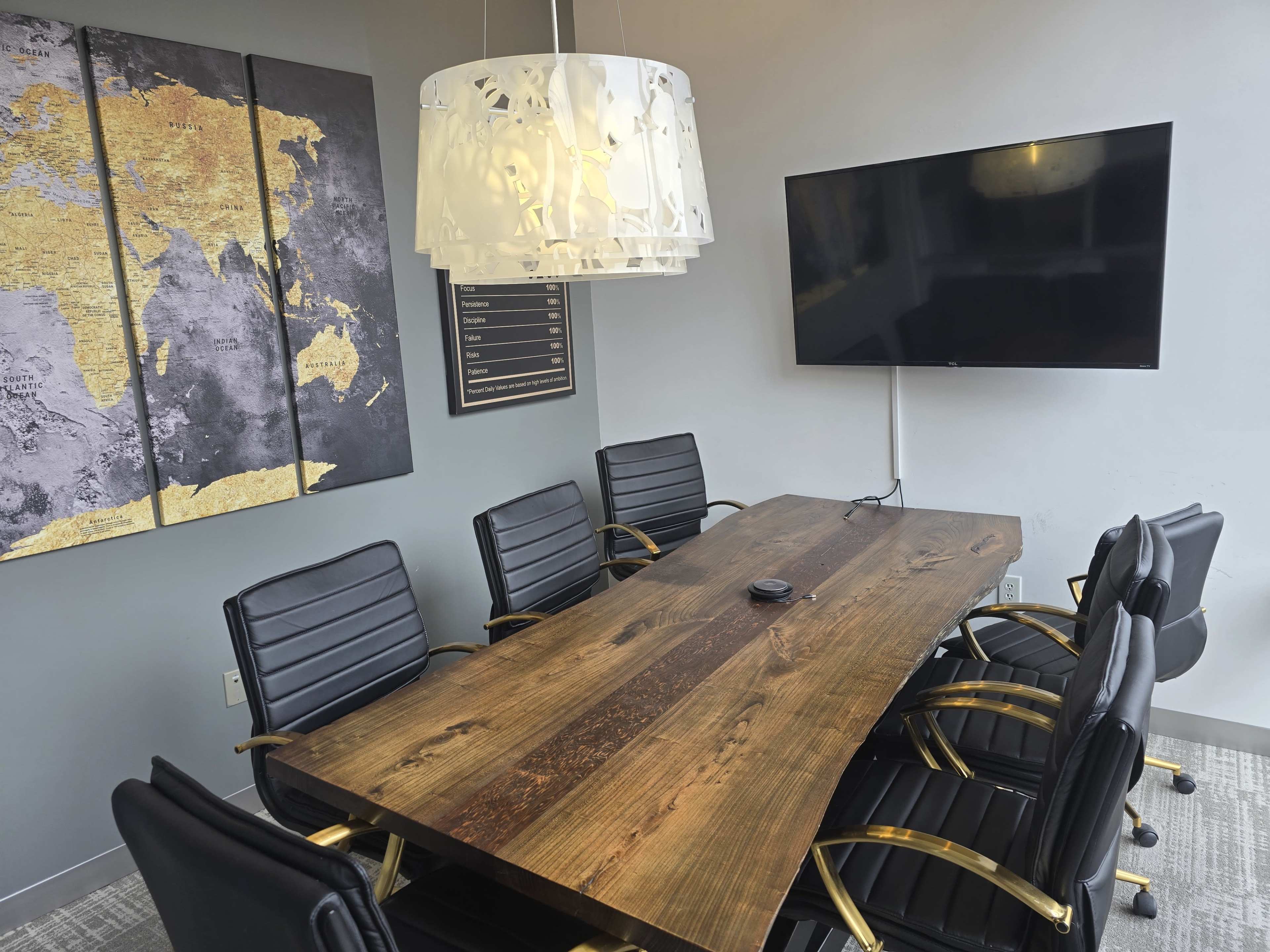 The image shows a conference room with a large wooden table surrounded by black chairs, a television on the wall, and a world map art piece in the background.