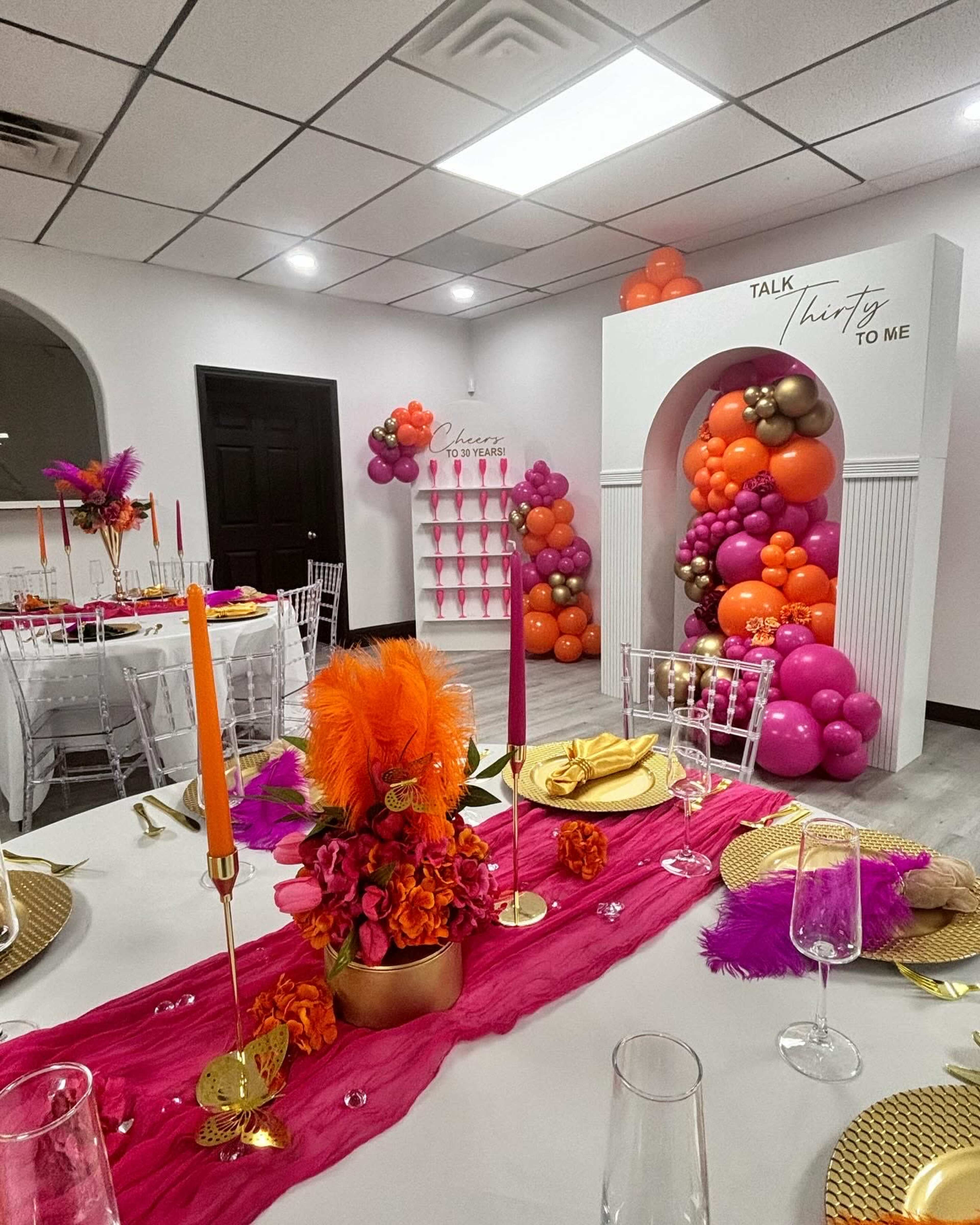 The image shows a decorated event space with tables set for a celebration, featuring bright balloons and colorful centerpieces.