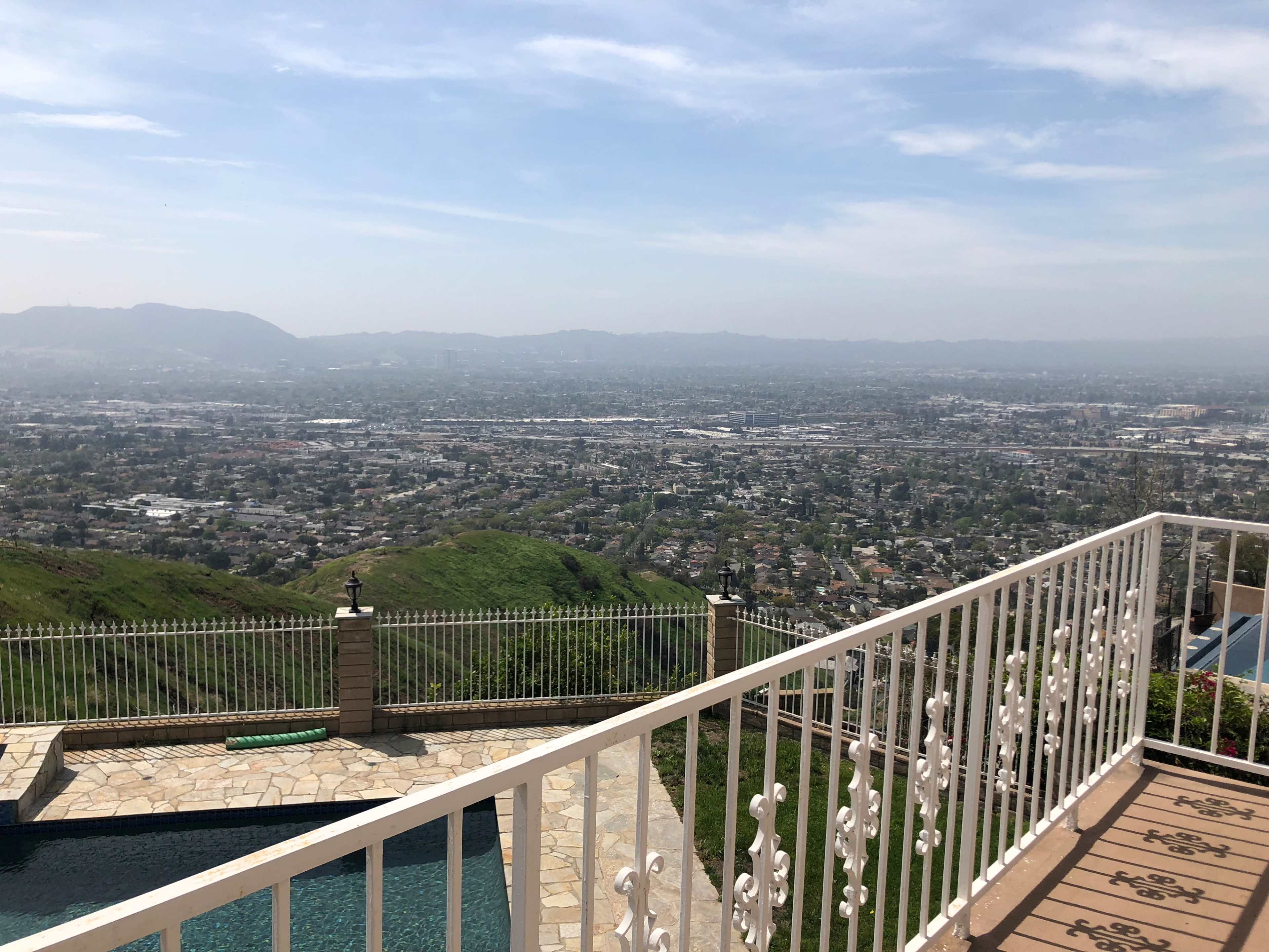 The image shows a panoramic view of a cityscape from an elevated terrace, with a swimming pool in the foreground and rolling hills in the distance.