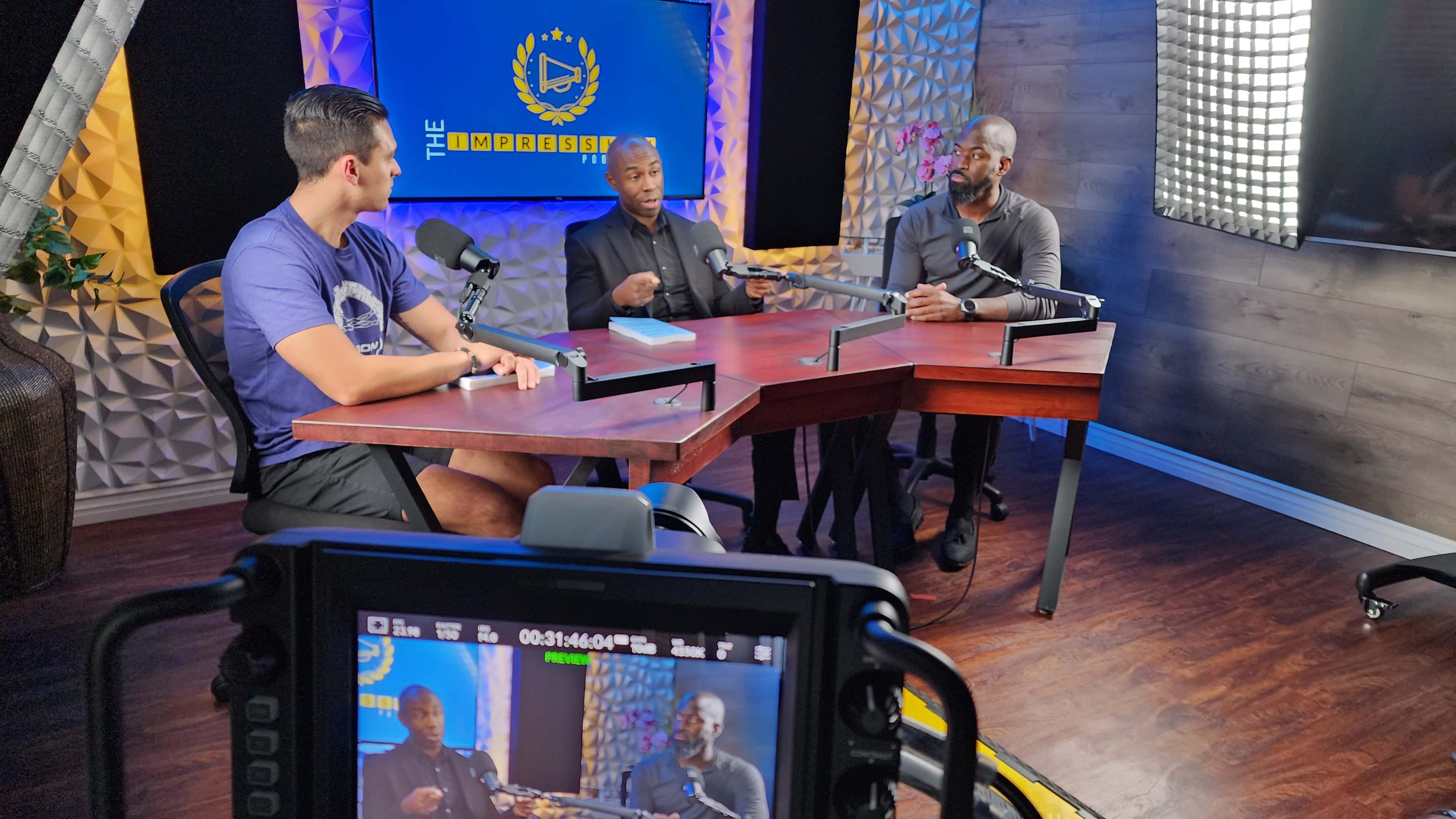 Three men are seated at a table in a studio setting, engaging in conversation while microphones are positioned in front of them.