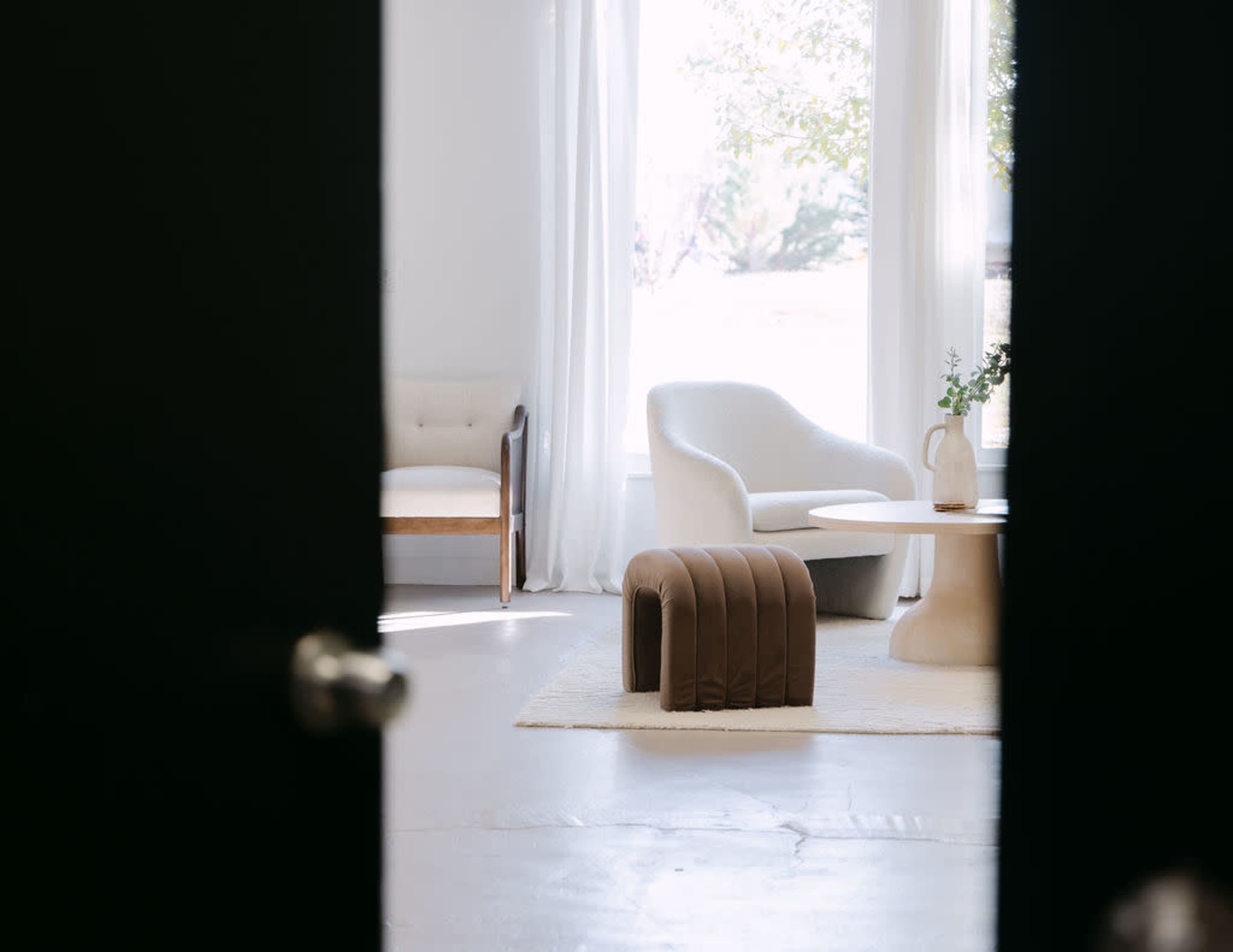 A modern interior space is visible through an open door, featuring a white armchair, a round table, a brown upholstered stool, and large windows letting in natural light.