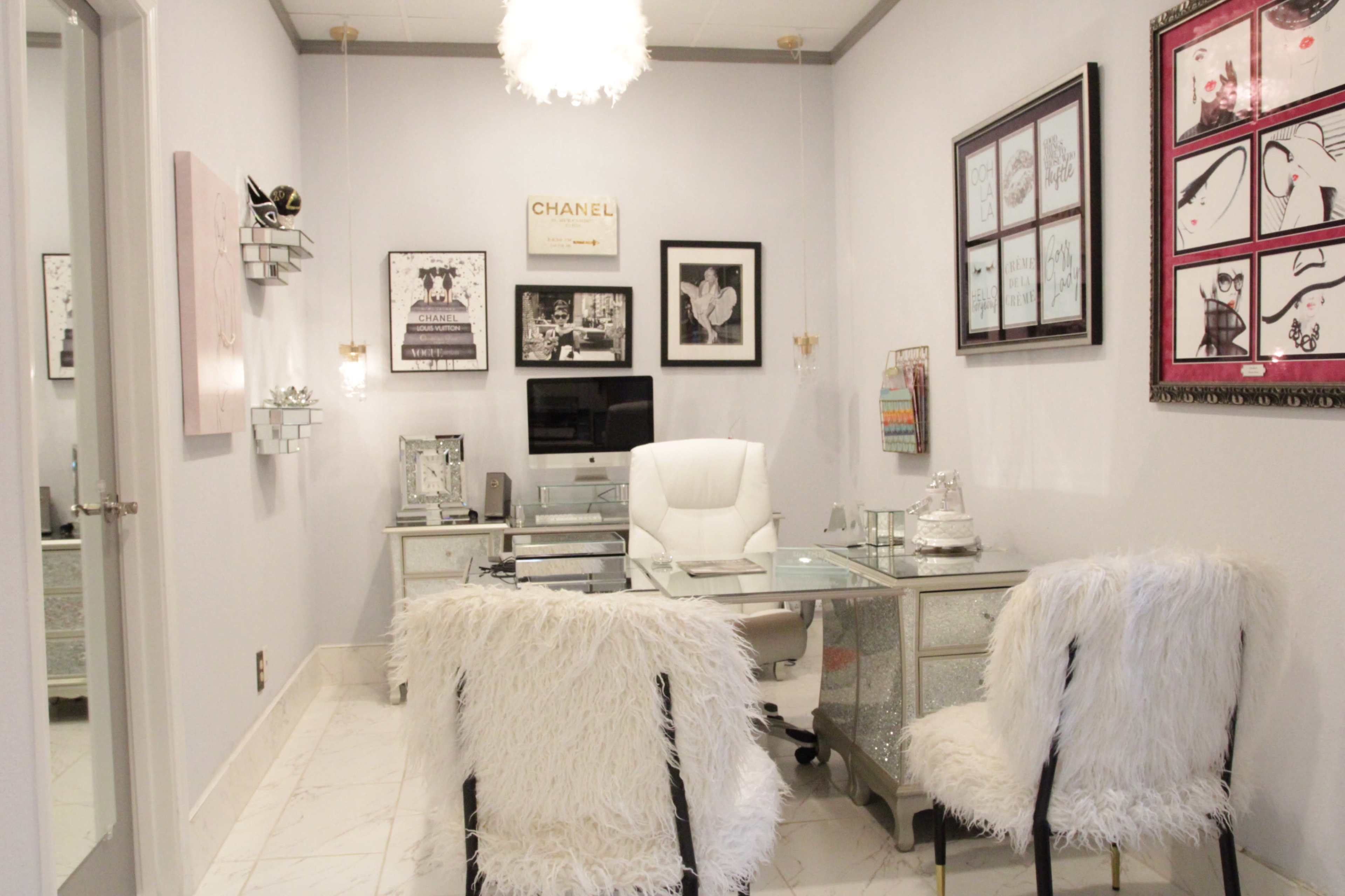 A stylish office features a glass desk, white chairs with furry upholstery, and framed artworks on light gray walls.