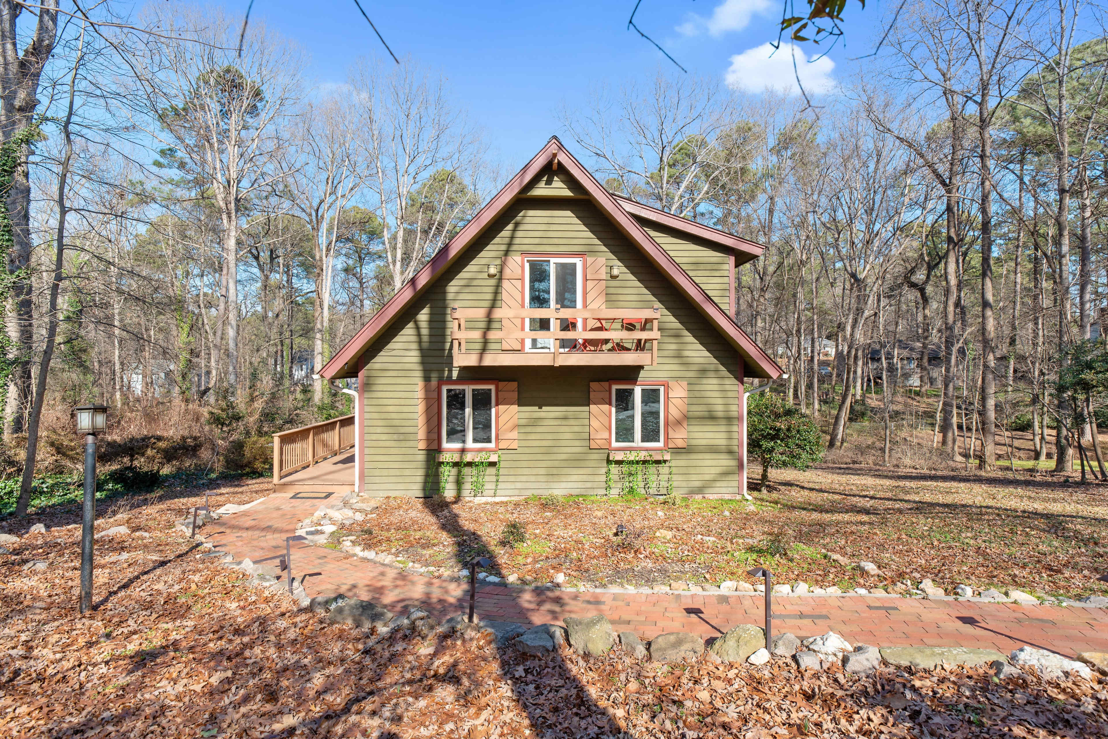 A green, two-story house with a pitched roof and wooden balcony overlooks a wooded area, surrounded by a brick pathway and scattered leaves.
