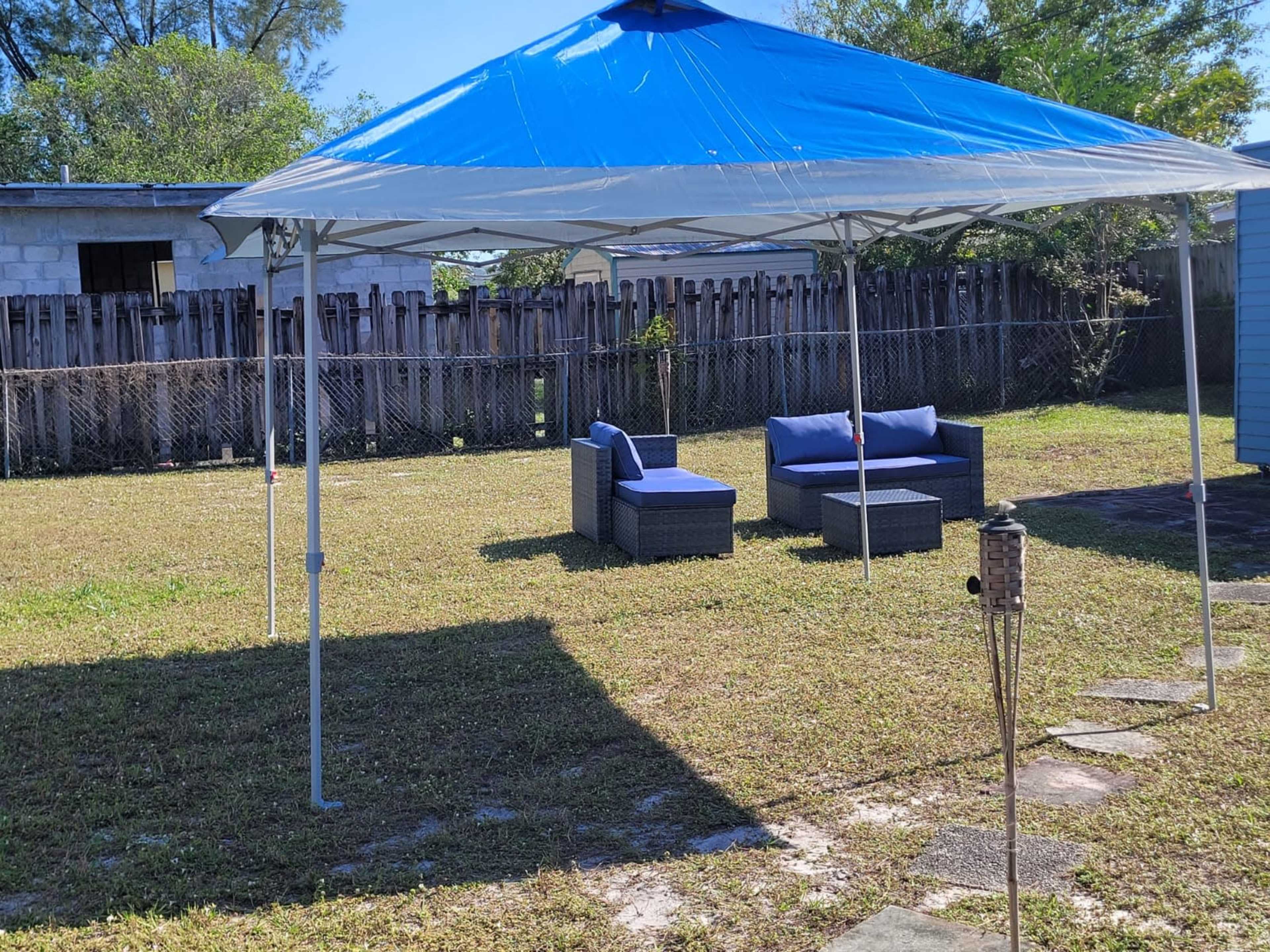 A blue and white canopy tent is set up in a grassy area with two cushioned chairs and a table beneath it.