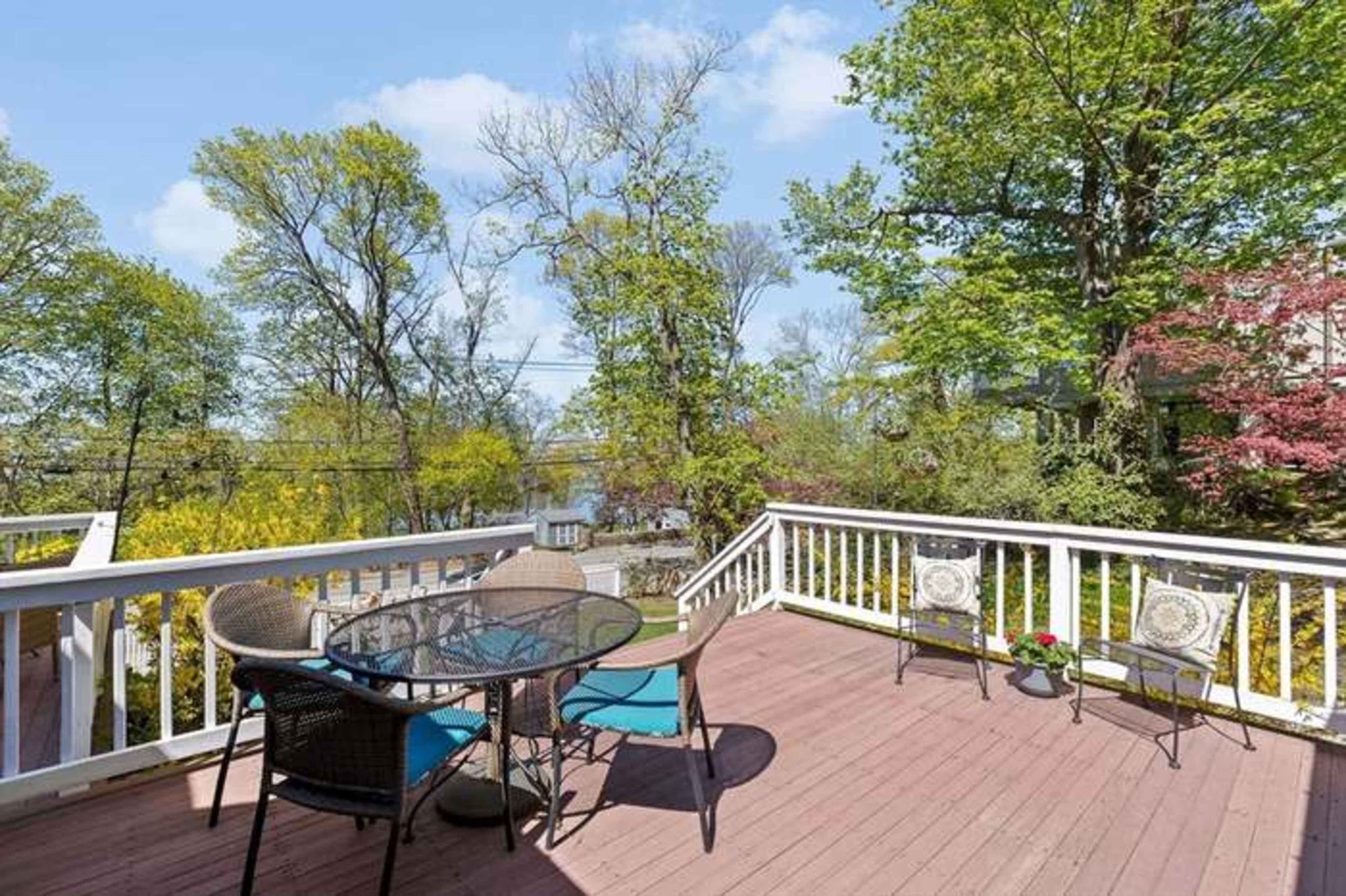 A wooden deck features a glass table and chairs surrounded by lush greenery and trees.