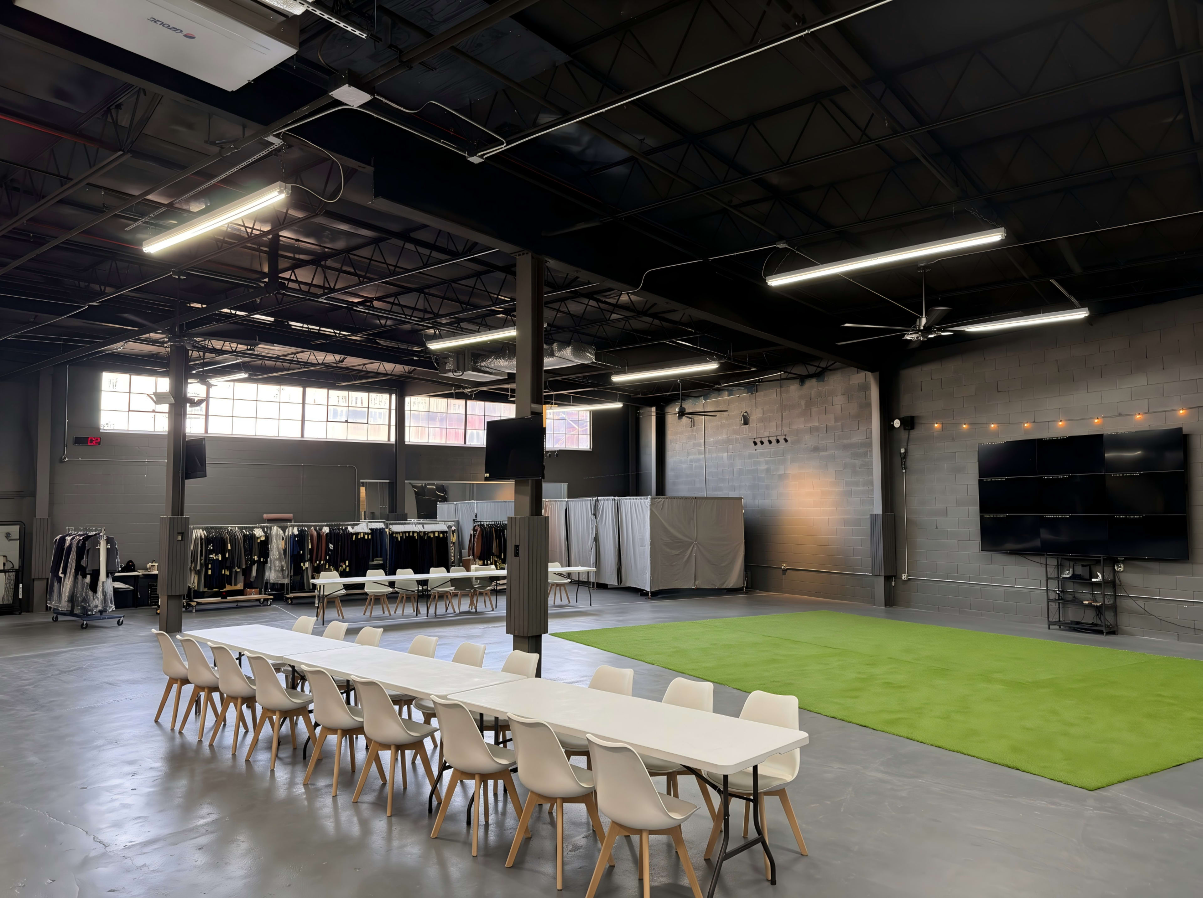 The image shows a spacious industrial-style room with a long table set for a meeting, a green carpeted area, and clothing racks along the walls.