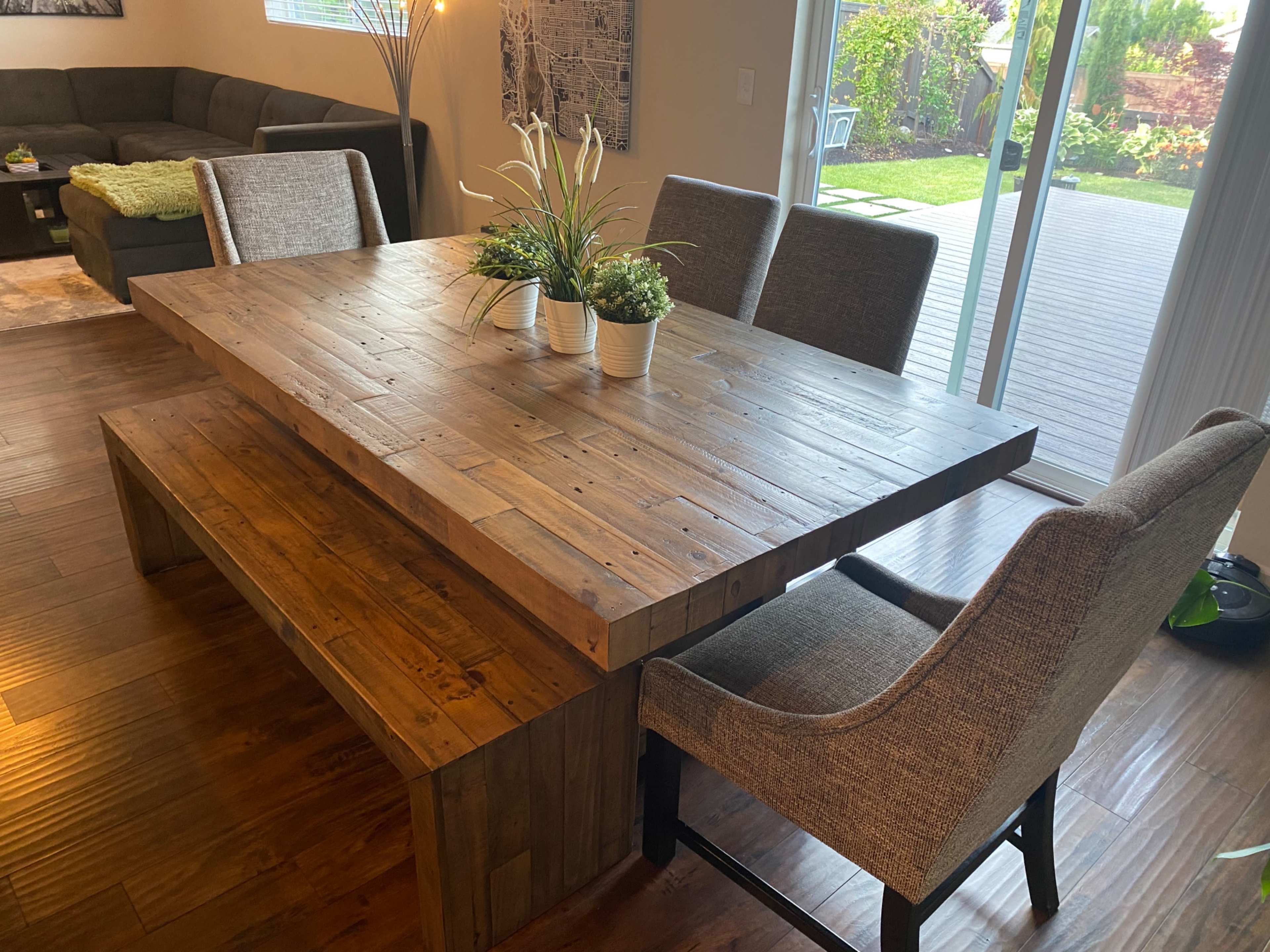 A wooden dining table with a bench on one side and four upholstered chairs arranged around it, set in a room with large windows overlooking a garden.