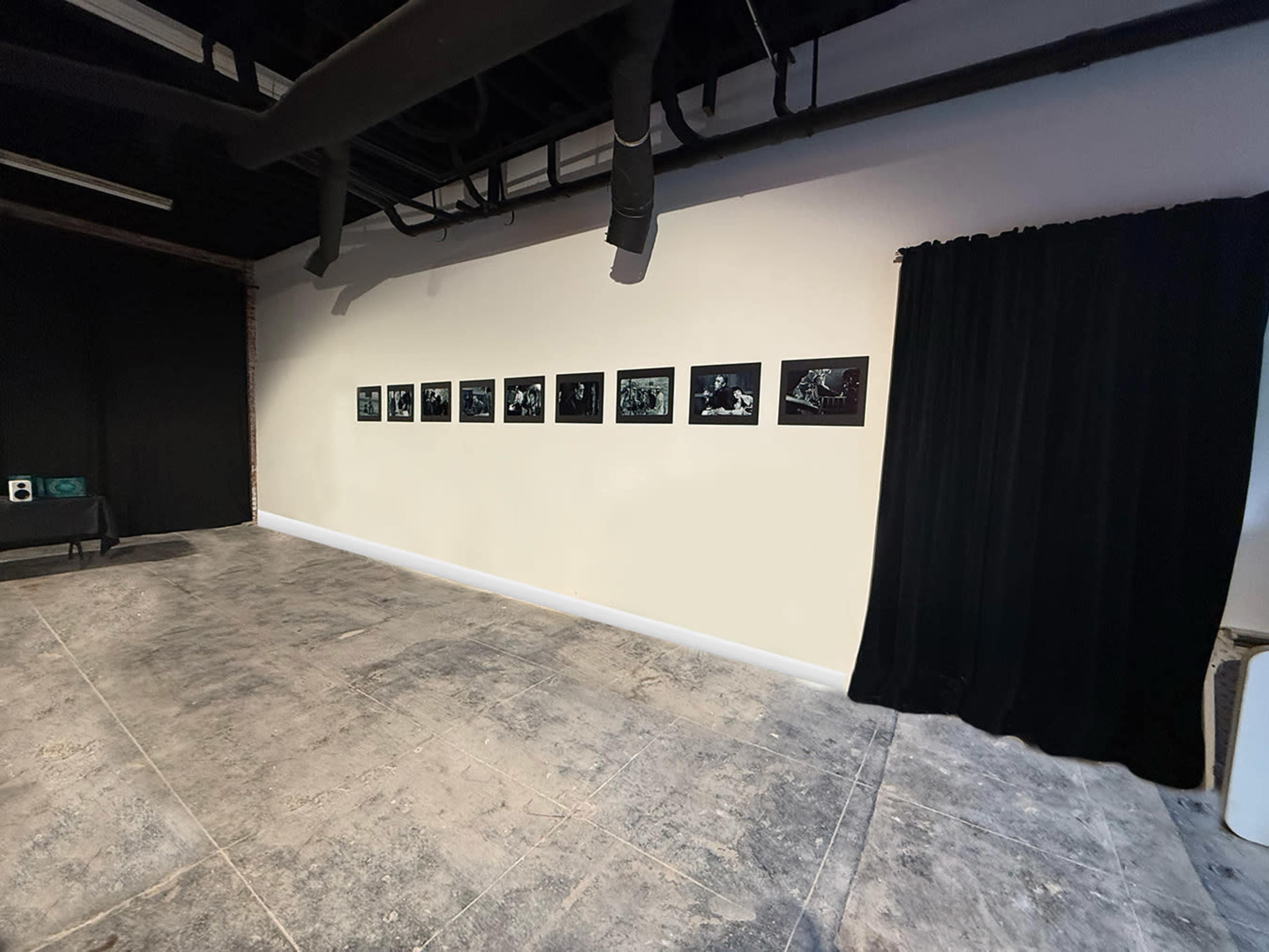 The image shows a gallery space featuring a line of eight black-and-white photographs displayed on a light-colored wall, with a dark curtain and bare floor in the background.