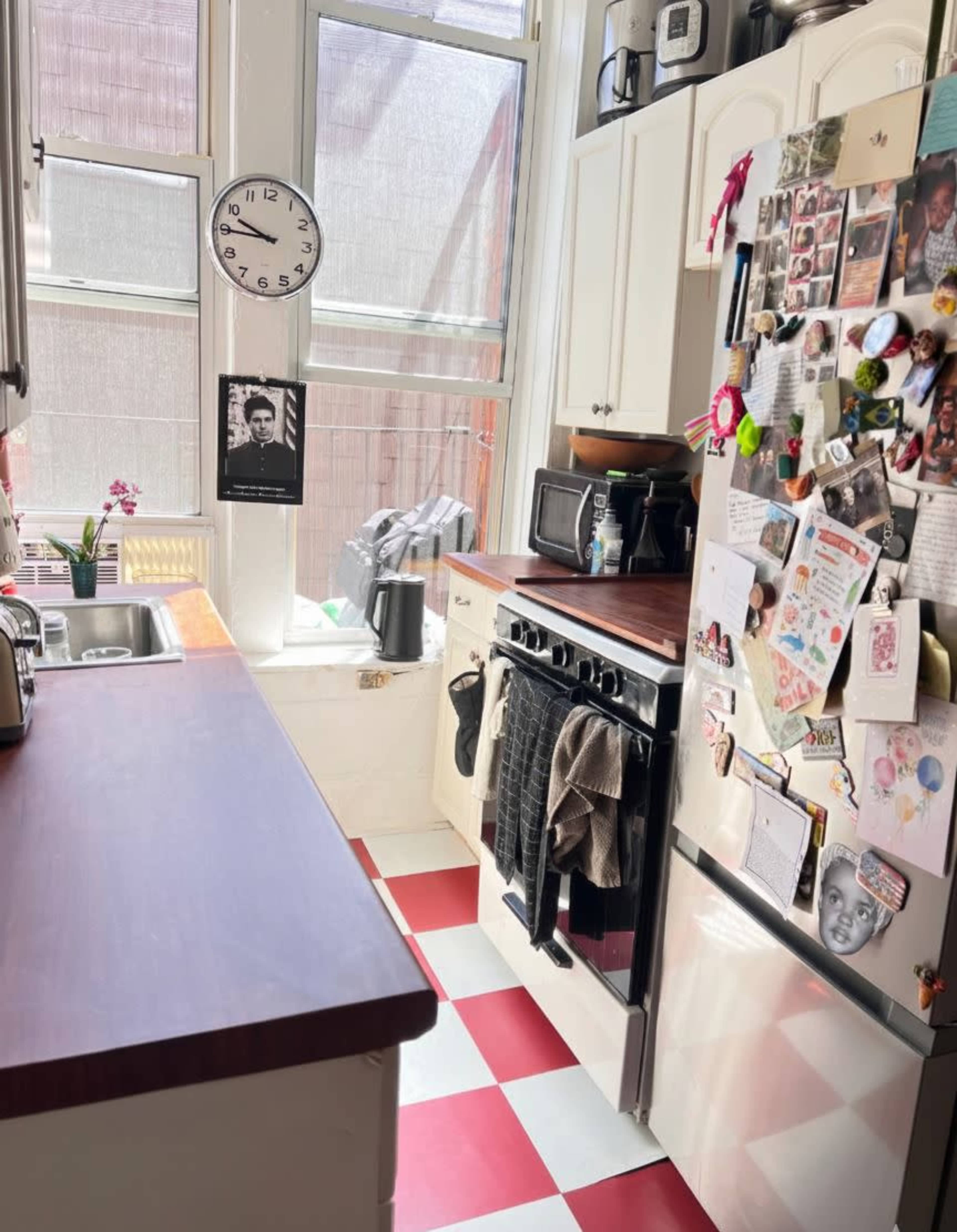 The image shows a small kitchen with a checkered red and white floor, a sink, a stove, a refrigerator covered in photographs and notes, and a window providing natural light.