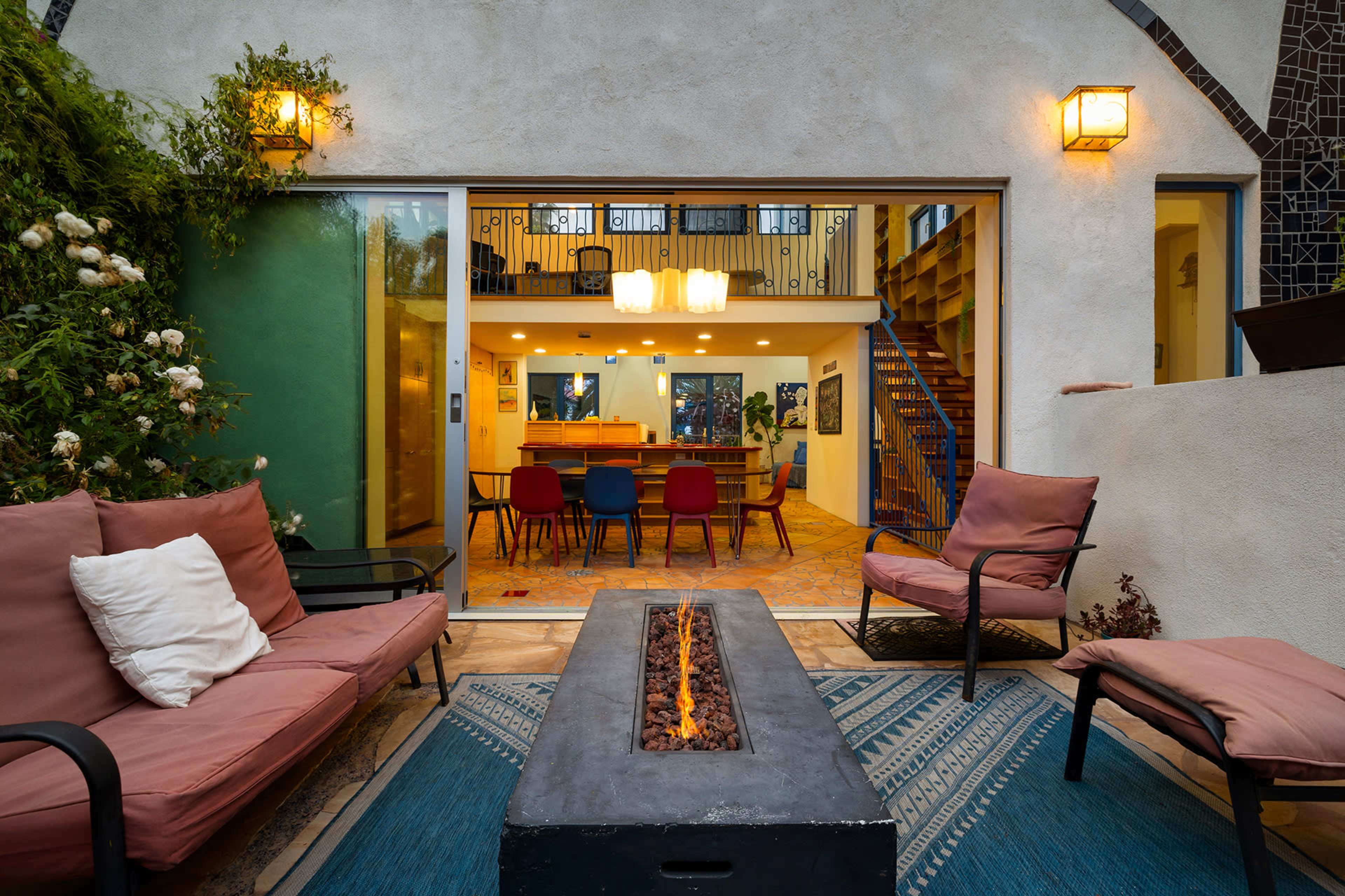 A cozy outdoor seating area features two chairs and a fire pit, with large glass doors opening to a brightly lit interior dining space.