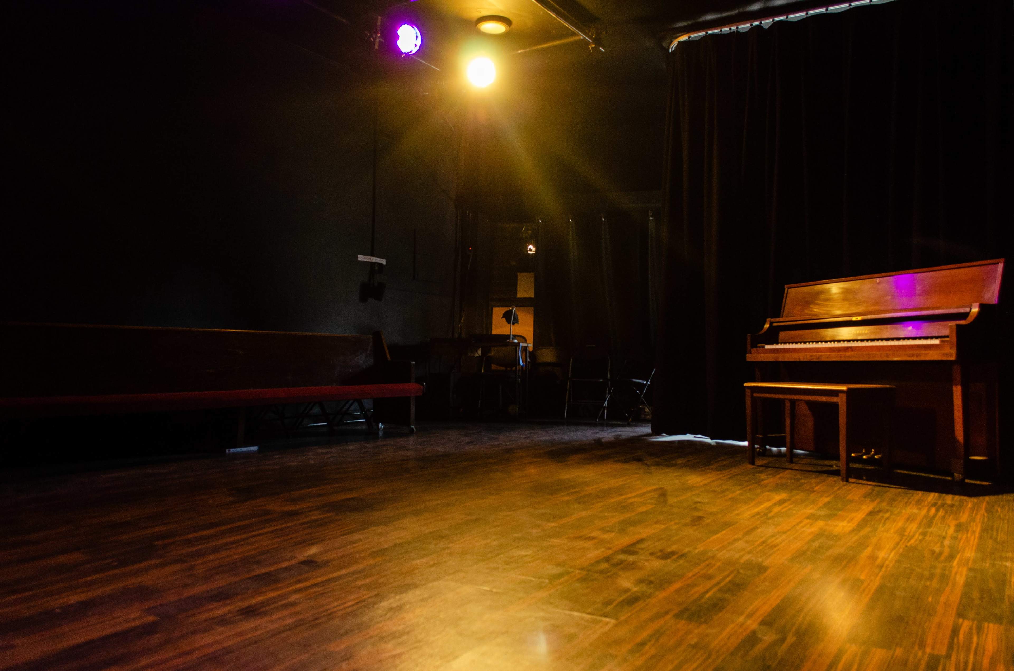 A dimly lit stage features a wooden piano and a few chairs in a small theater setting.