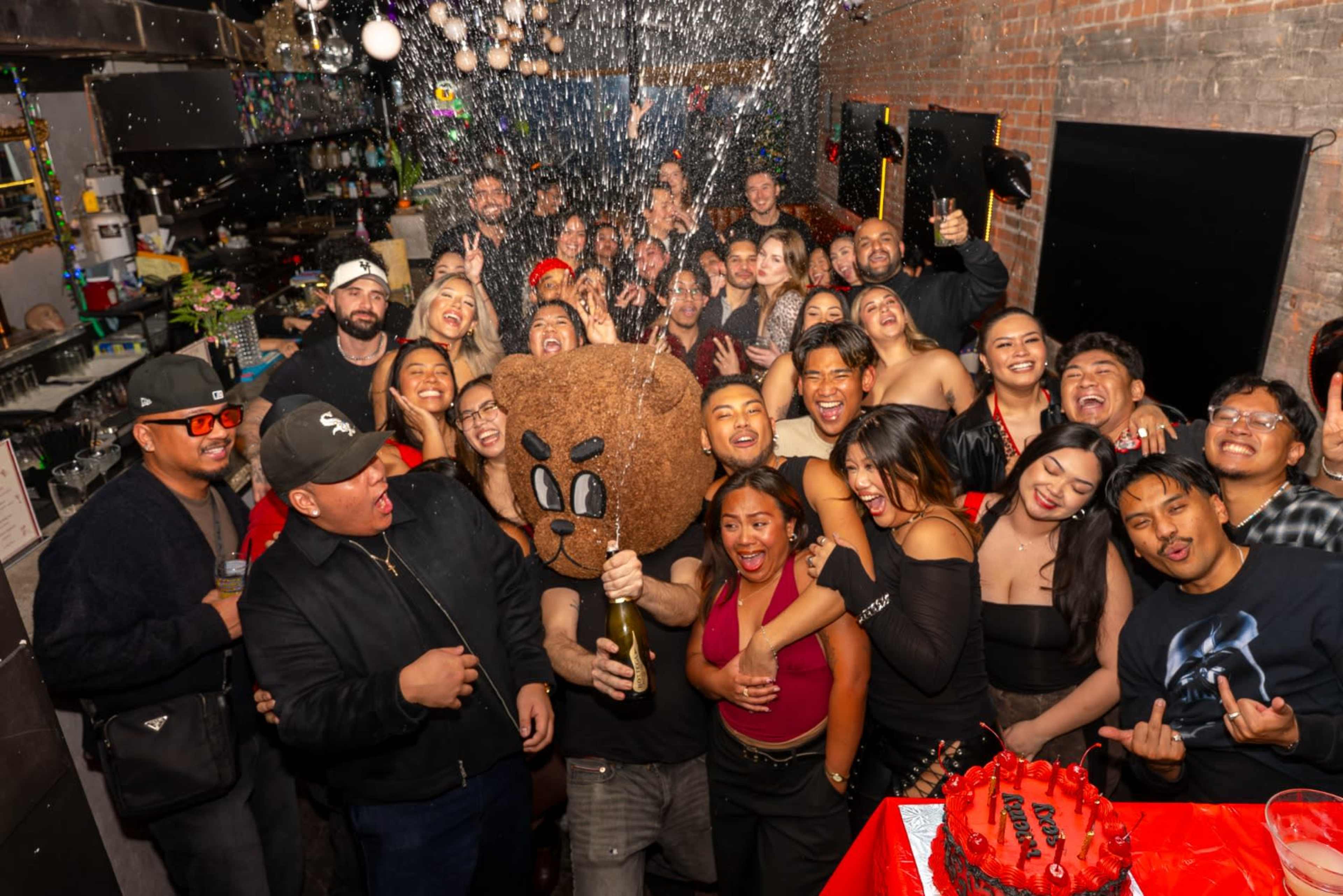 A large group of people is celebrating indoors, with one person wearing a large bear-head costume while others are posing and cheering with drinks.