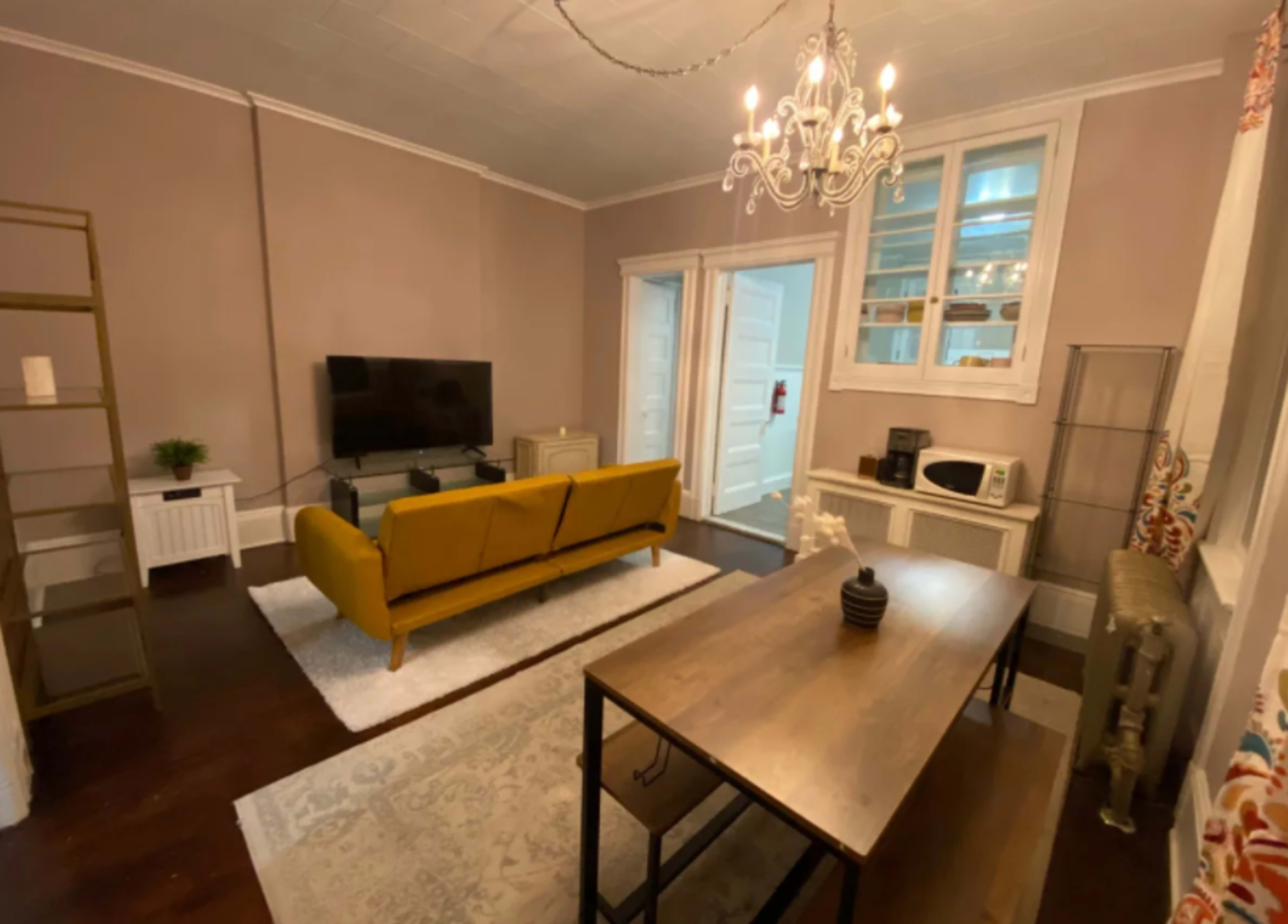 A living room with a yellow couch, a television mounted on the wall, a wooden table, and a microwave on a shelf, all beneath a chandelier.