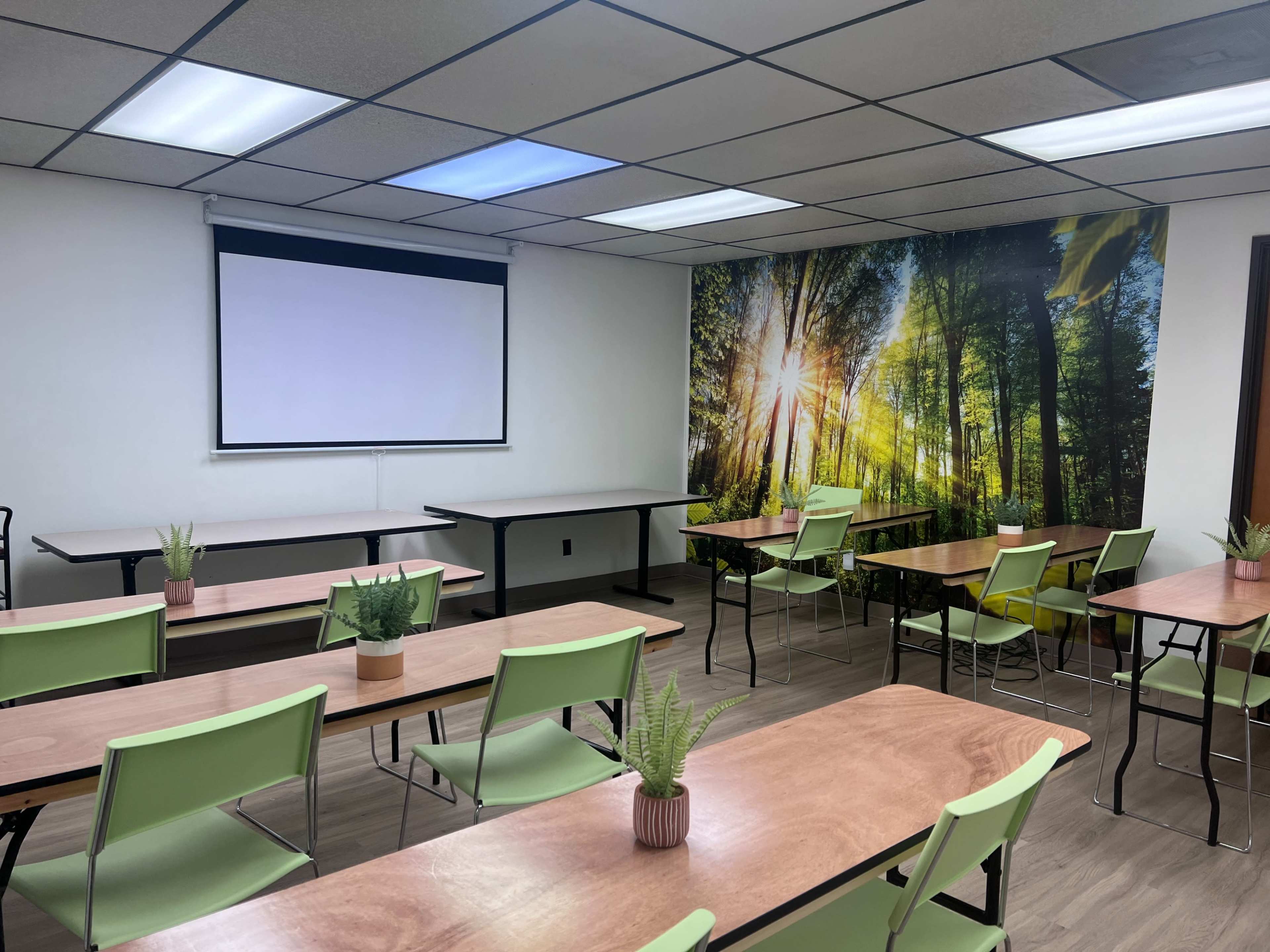 A classroom with tables and chairs arranged for a meeting, featuring a projection screen and a wall mural of a forest scene.