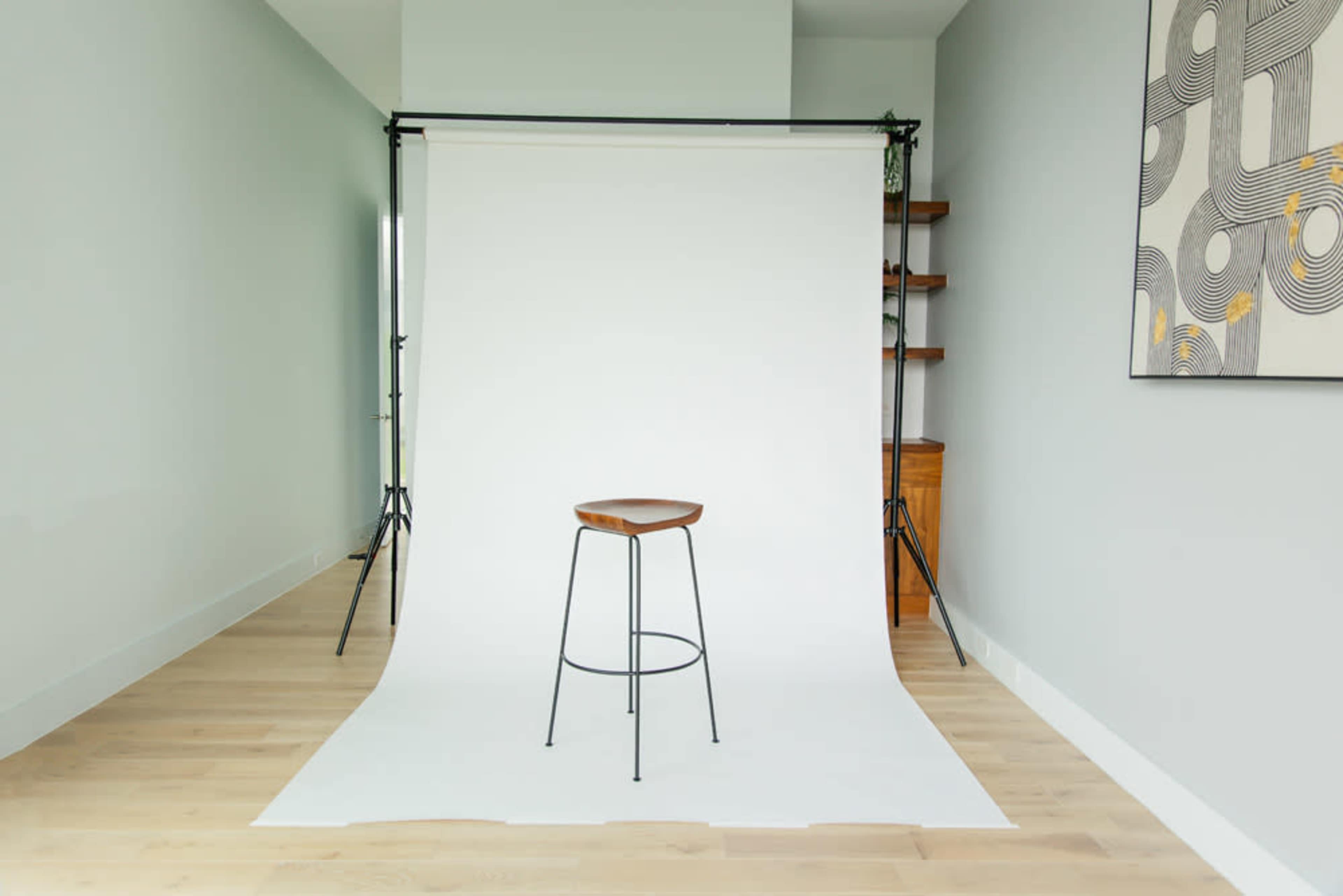 The image shows a minimalistic studio setup with a single wooden stool in front of a white backdrop and light-colored wooden flooring.