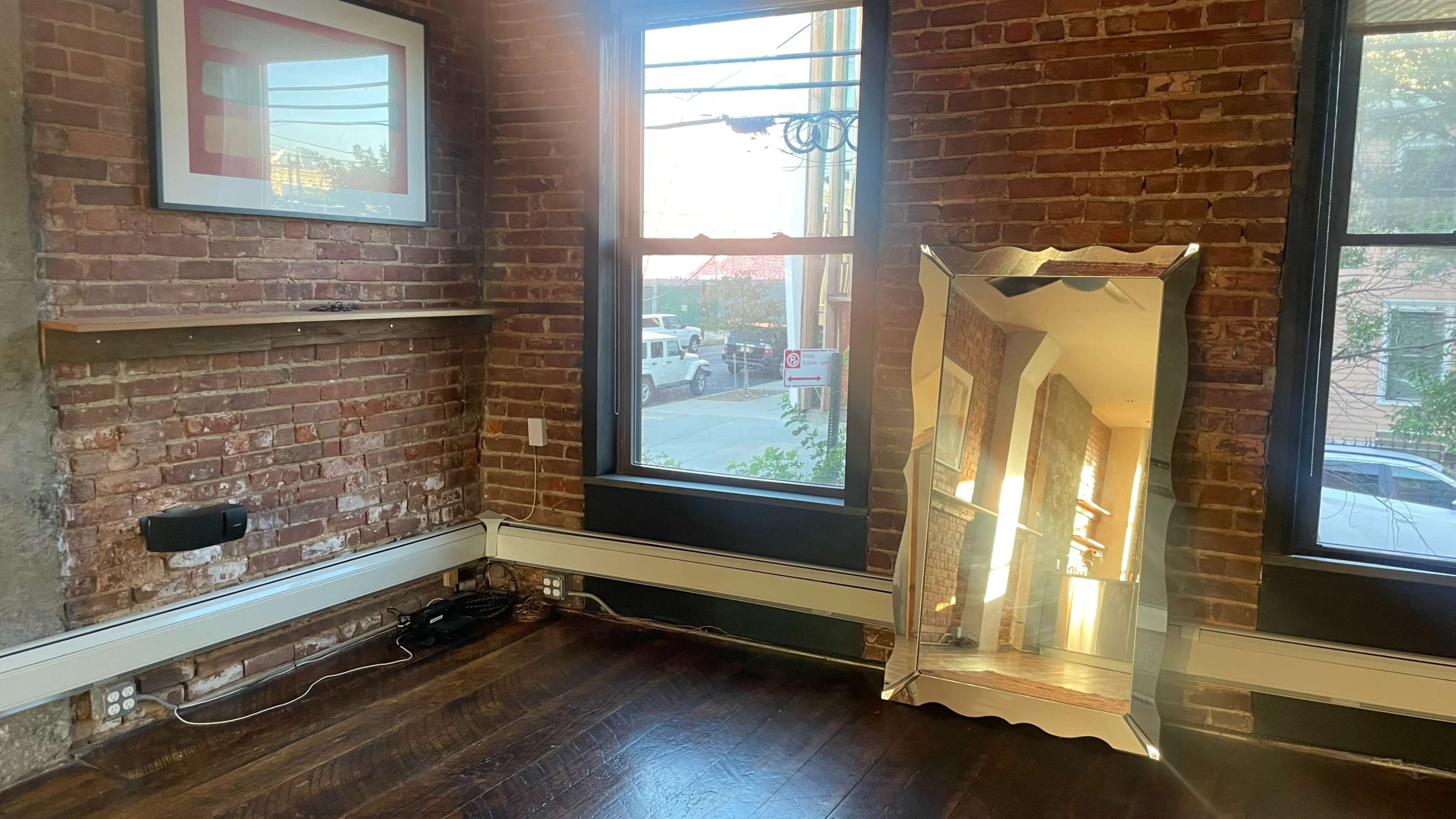 A room with exposed brick walls, a large mirror reflecting light, and two windows overlooking a street.