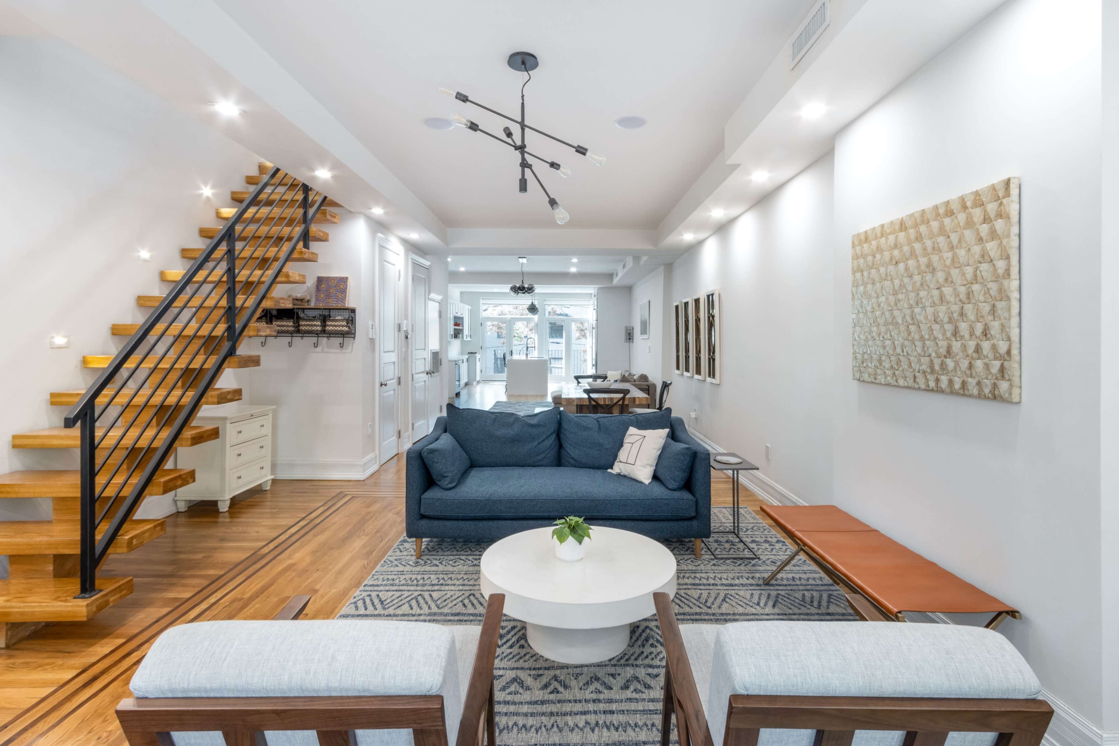 The image shows a modern living room with a blue sofa, two light-colored armchairs, a round coffee table, and a wooden staircase leading to an upper level.