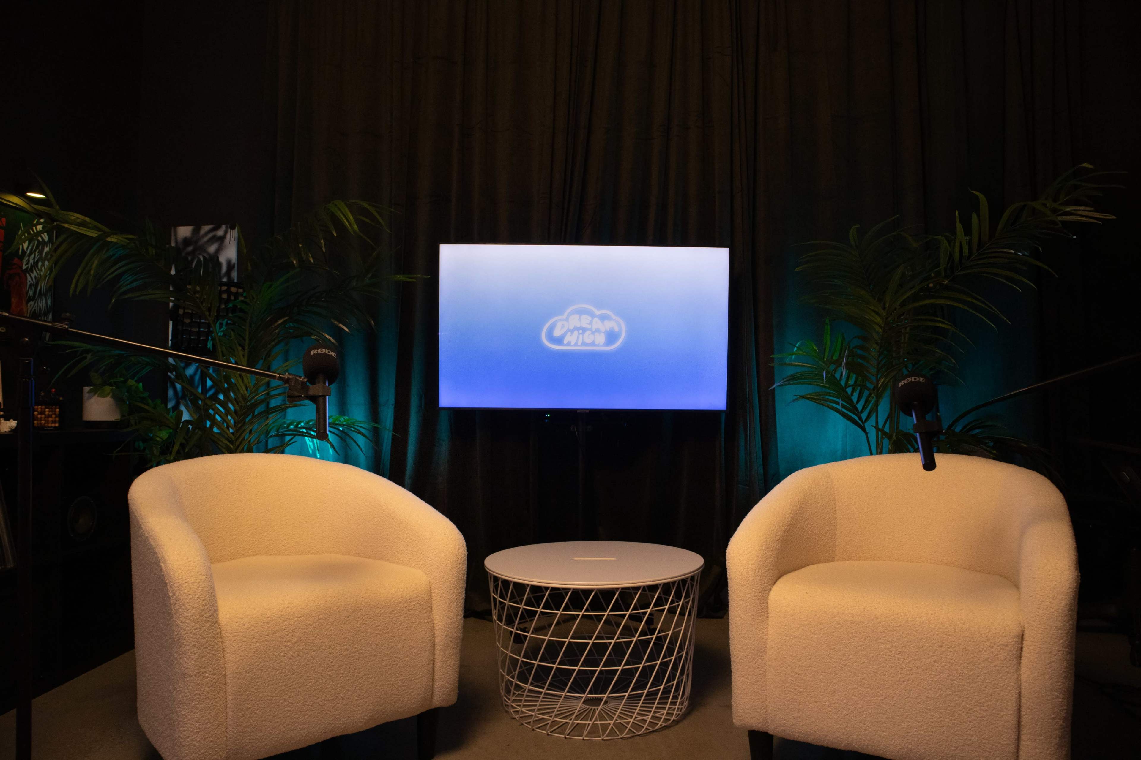 The image shows a modern viewing area with two white armchairs, a round table, and a television displaying a logo, all set against a dark backdrop with green lighting and indoor plants.