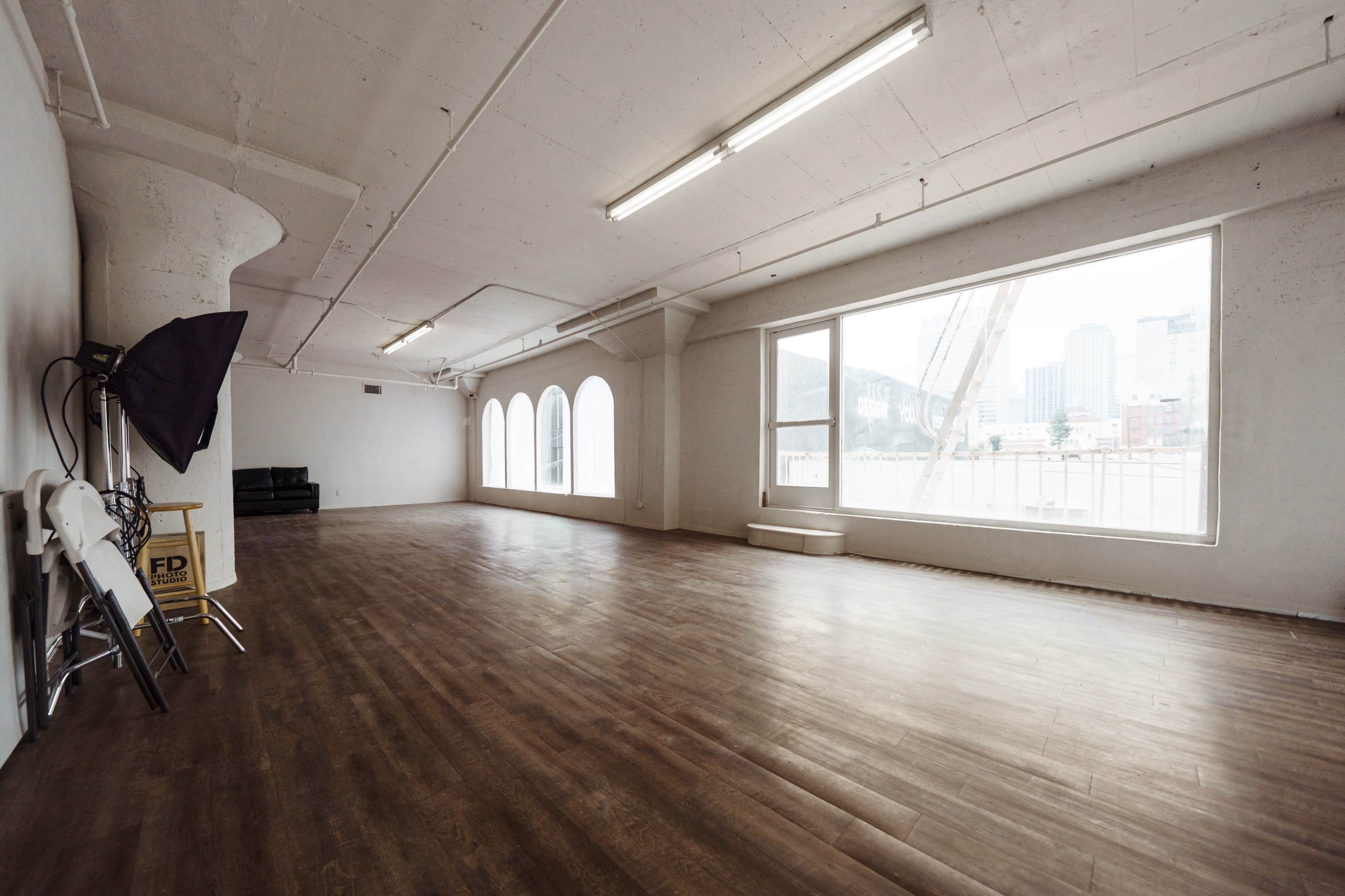 An empty, spacious room with large windows and a wooden floor, featuring minimal furniture and architectural details.