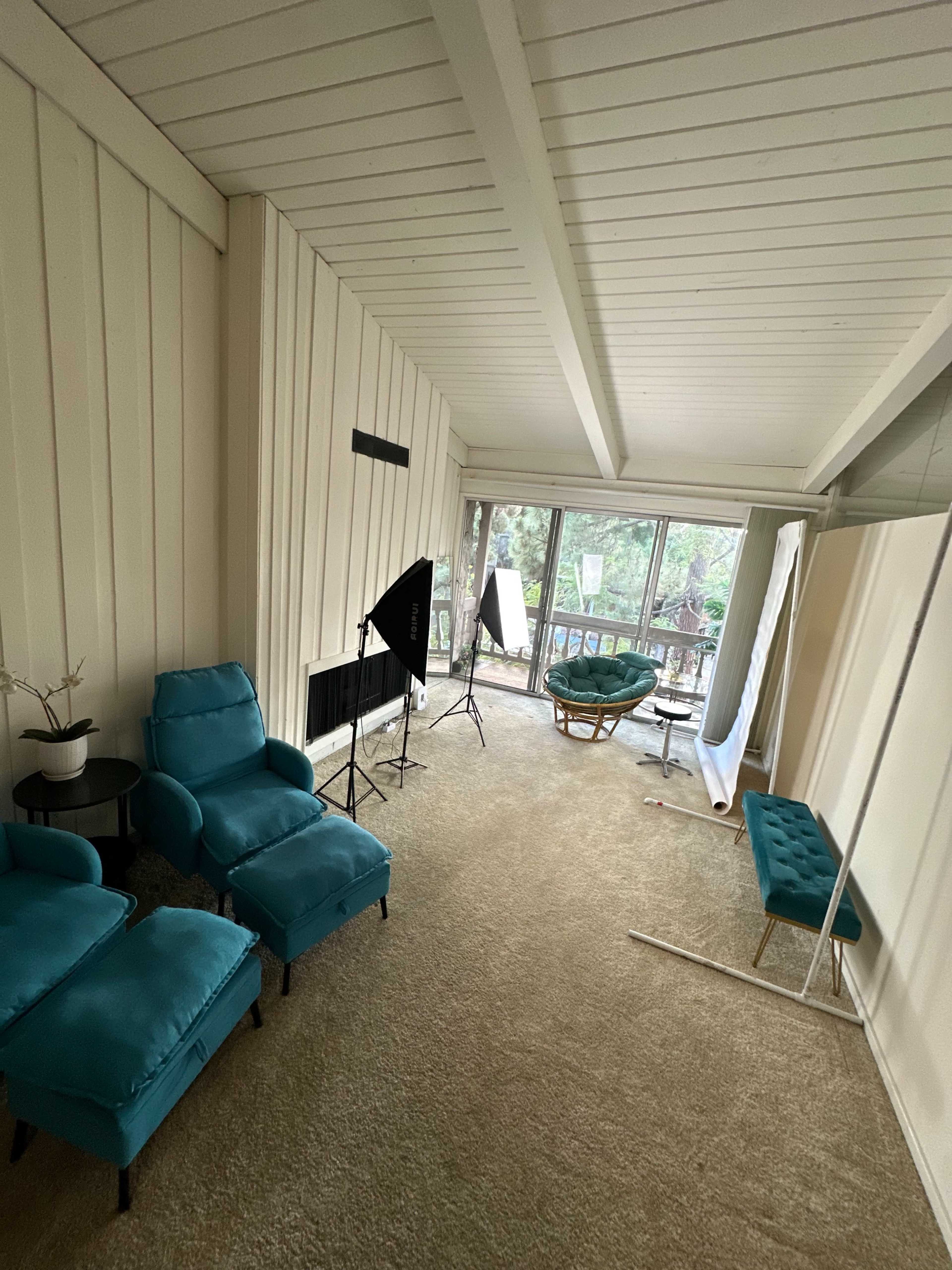 The image shows a bright, airy room with large windows, featuring turquoise furniture, a light-colored carpet, and photography equipment set up in one corner.