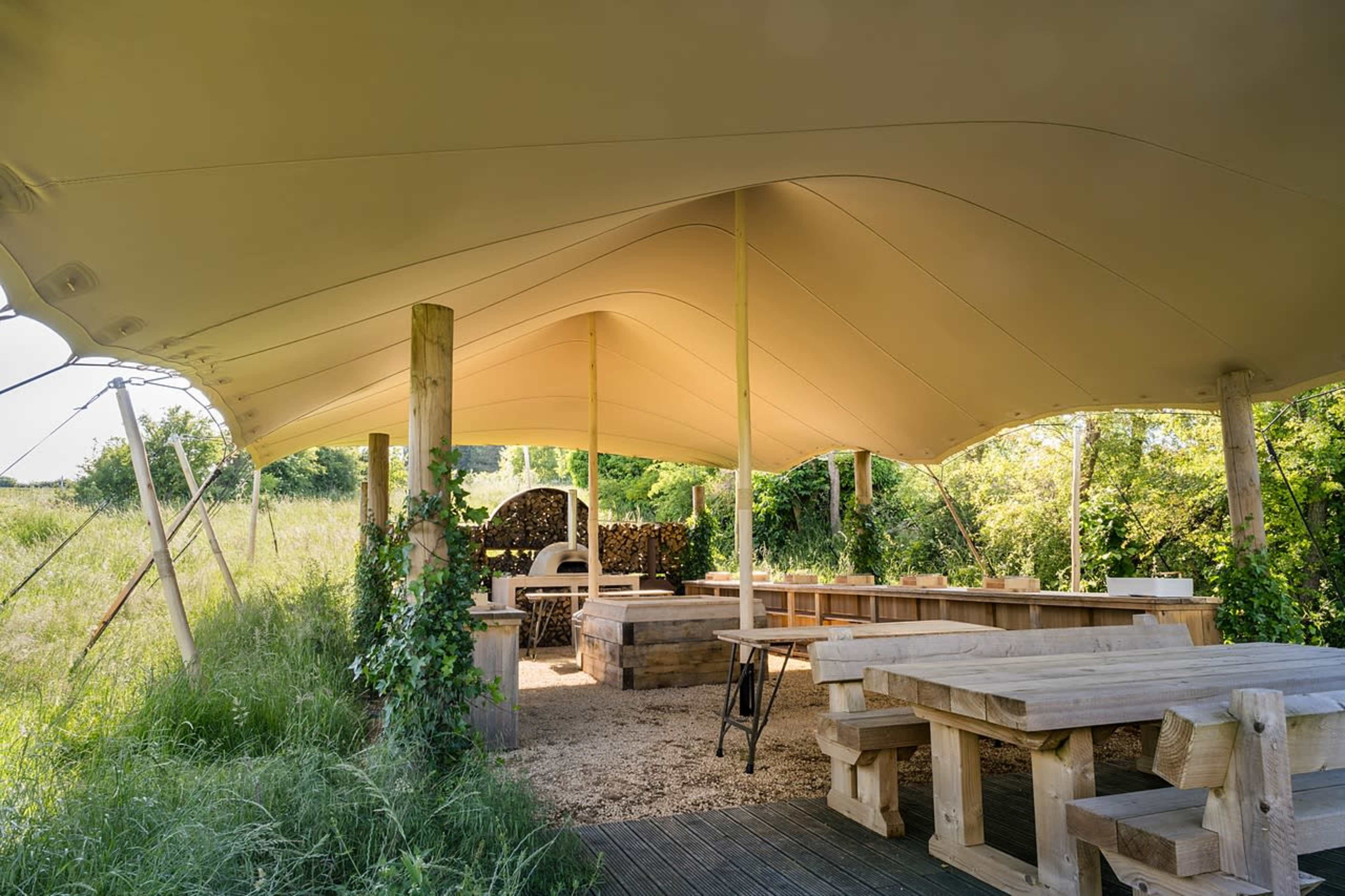 The scene features an outdoor gazebo with a large fabric canopy, wooden tables, and a stacked woodpile surrounded by greenery.