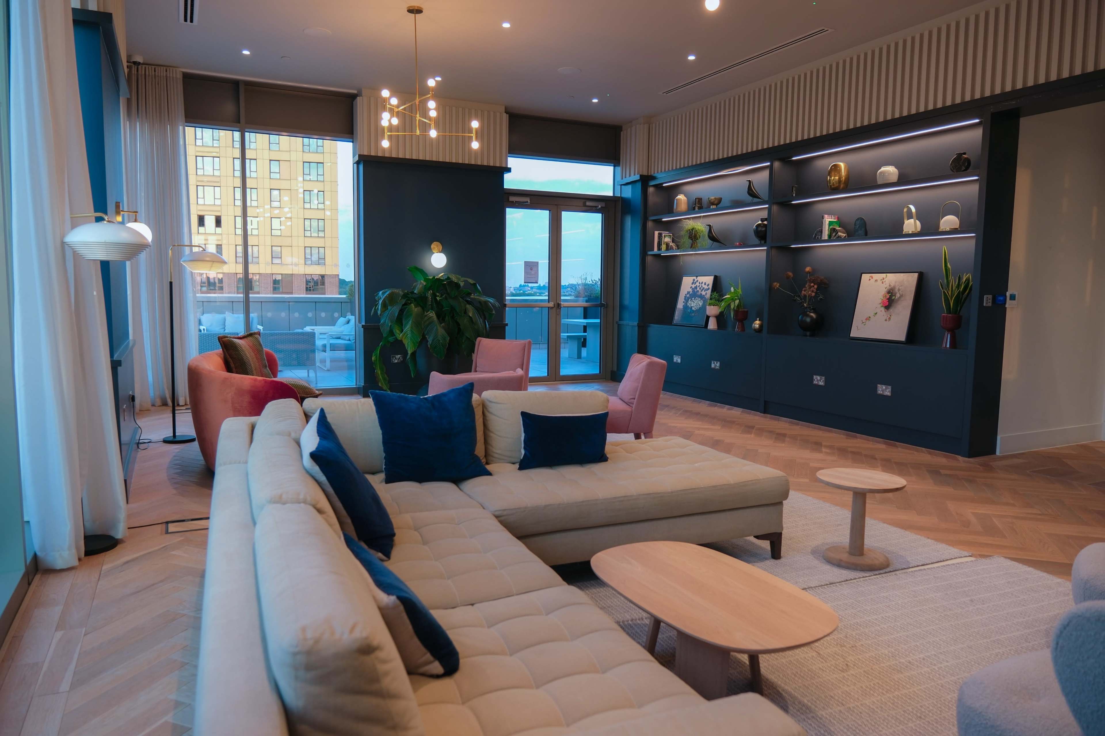 A modern living space featuring a beige sectional sofa, pink accent chairs, a round coffee table, and shelves displaying decorative items against a blue wall.
