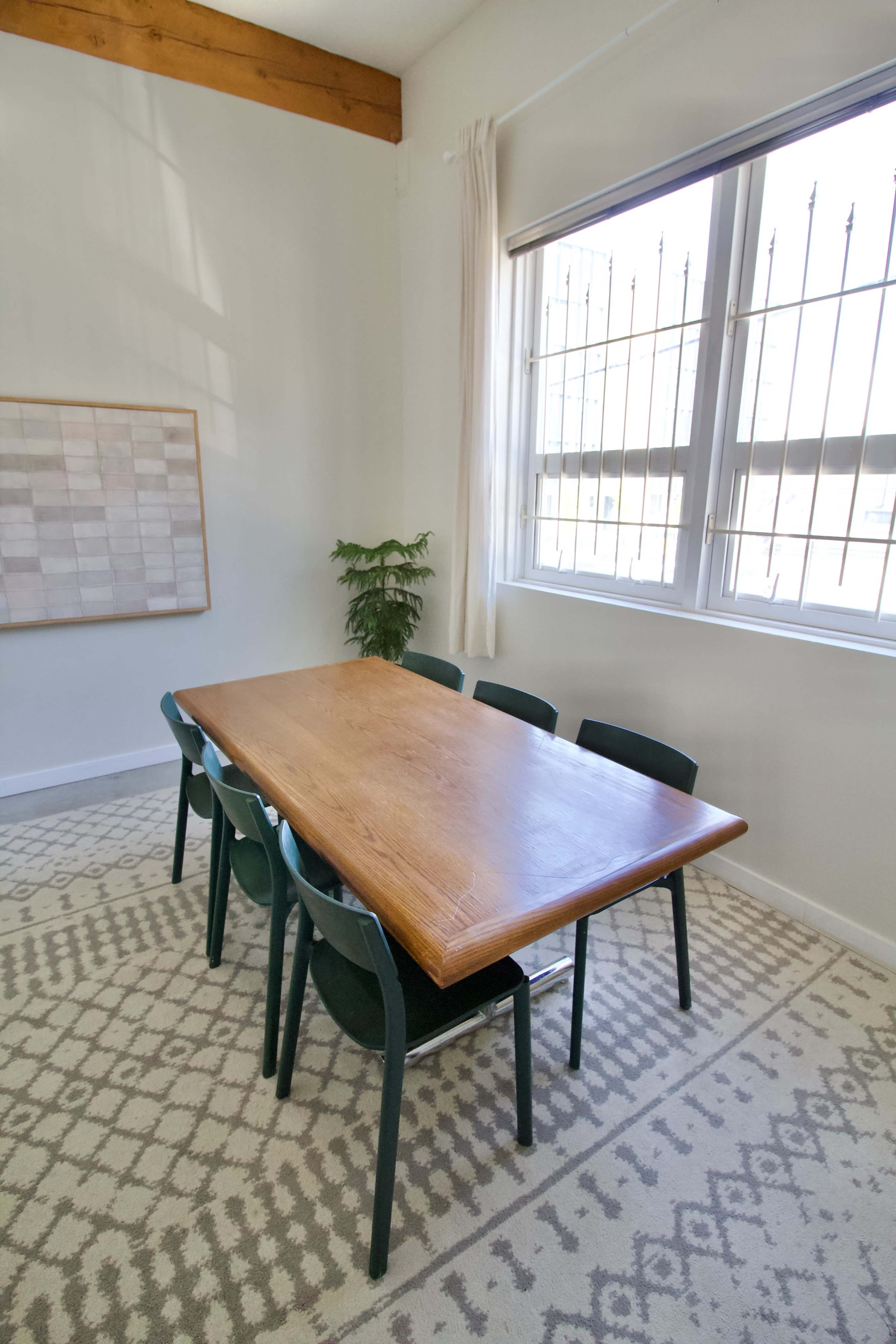 A wooden dining table surrounded by six green chairs is positioned in a well-lit room with a patterned rug and a small potted plant near the window.