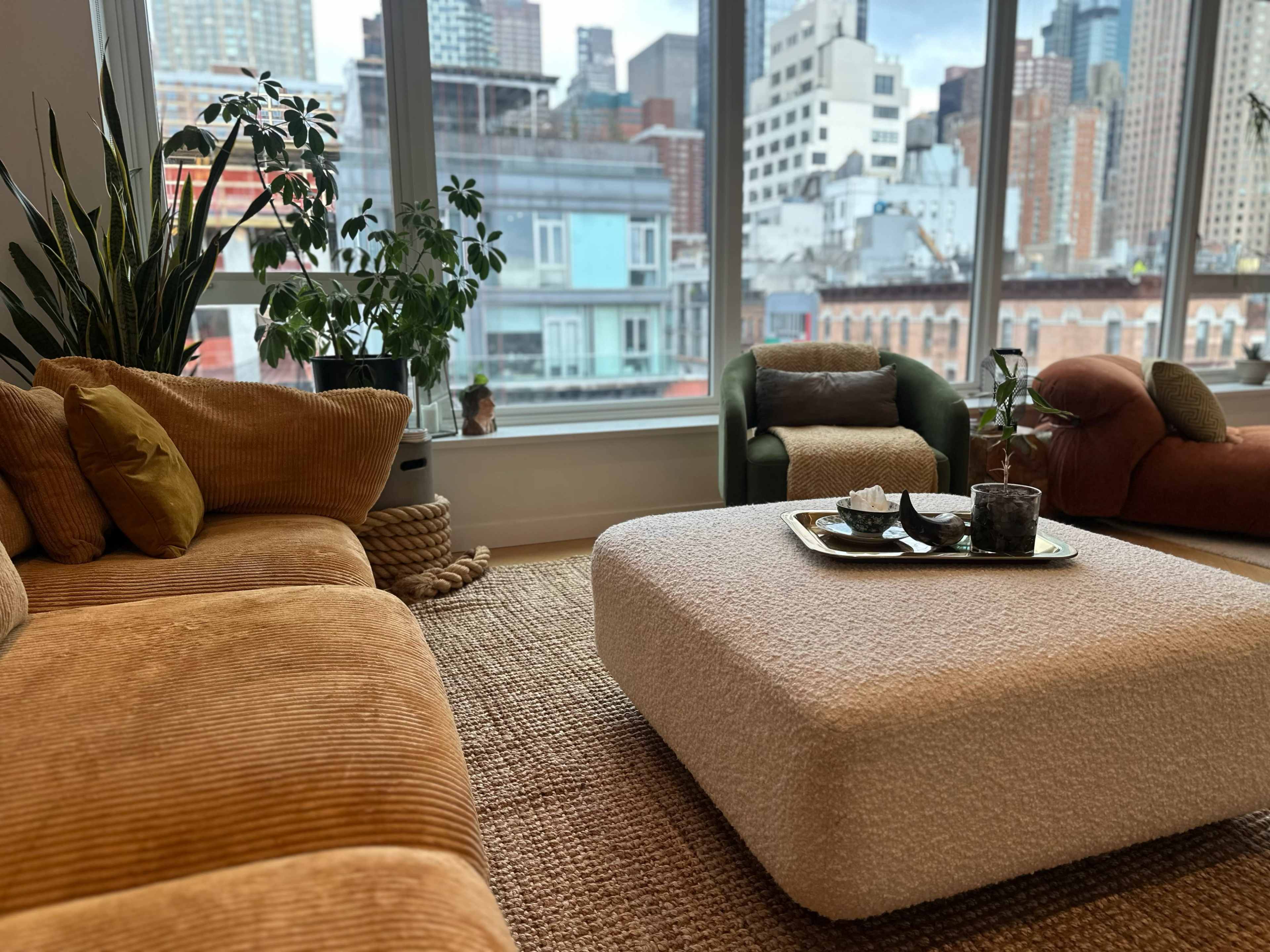 The image shows a modern living room with a large, plush sectional sofa, a cozy coffee table with decorative items, and large windows overlooking a city skyline.