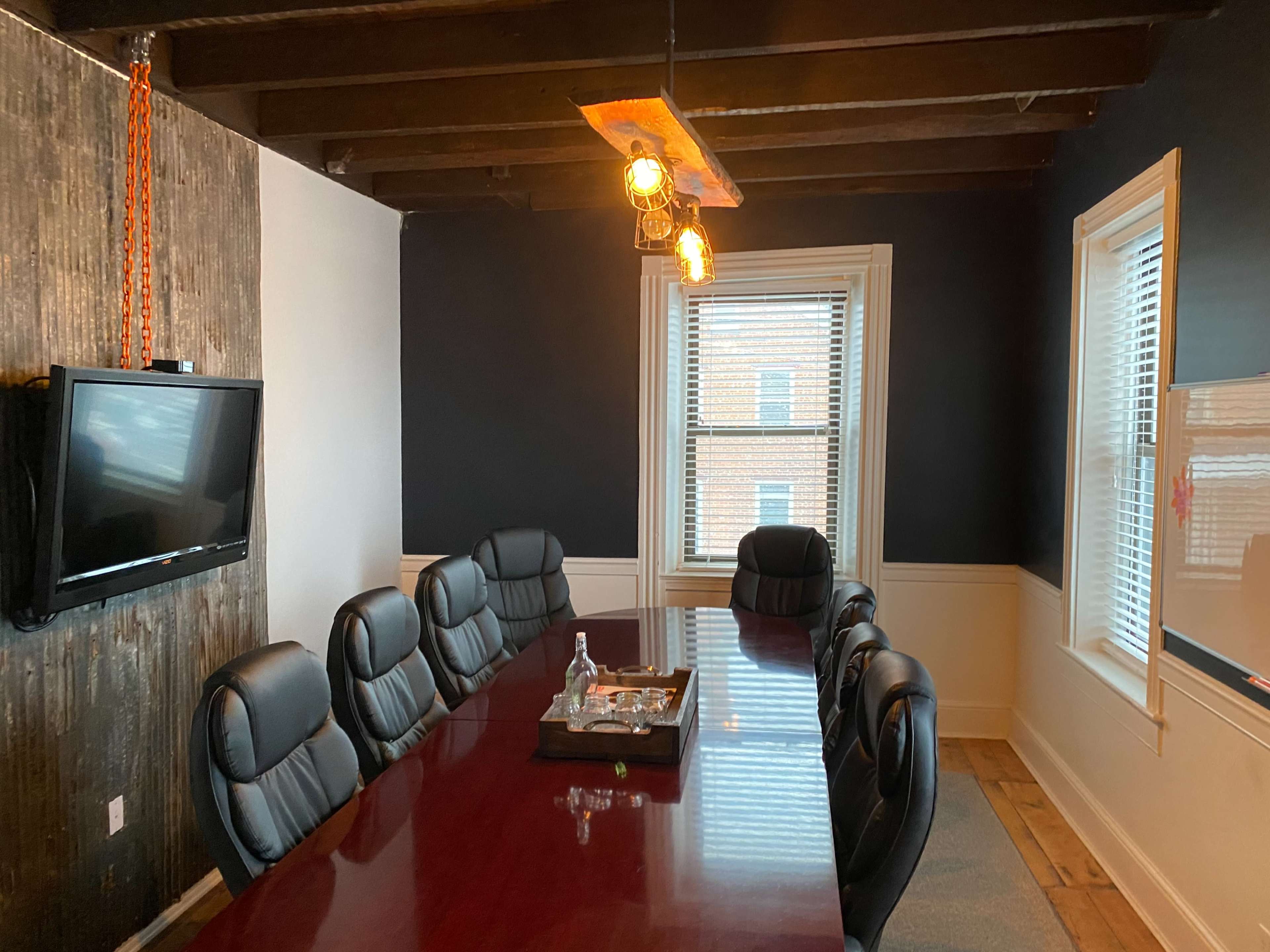The image shows a conference room with a long wooden table, black leather chairs, a television mounted on the wall, and large windows with blinds.