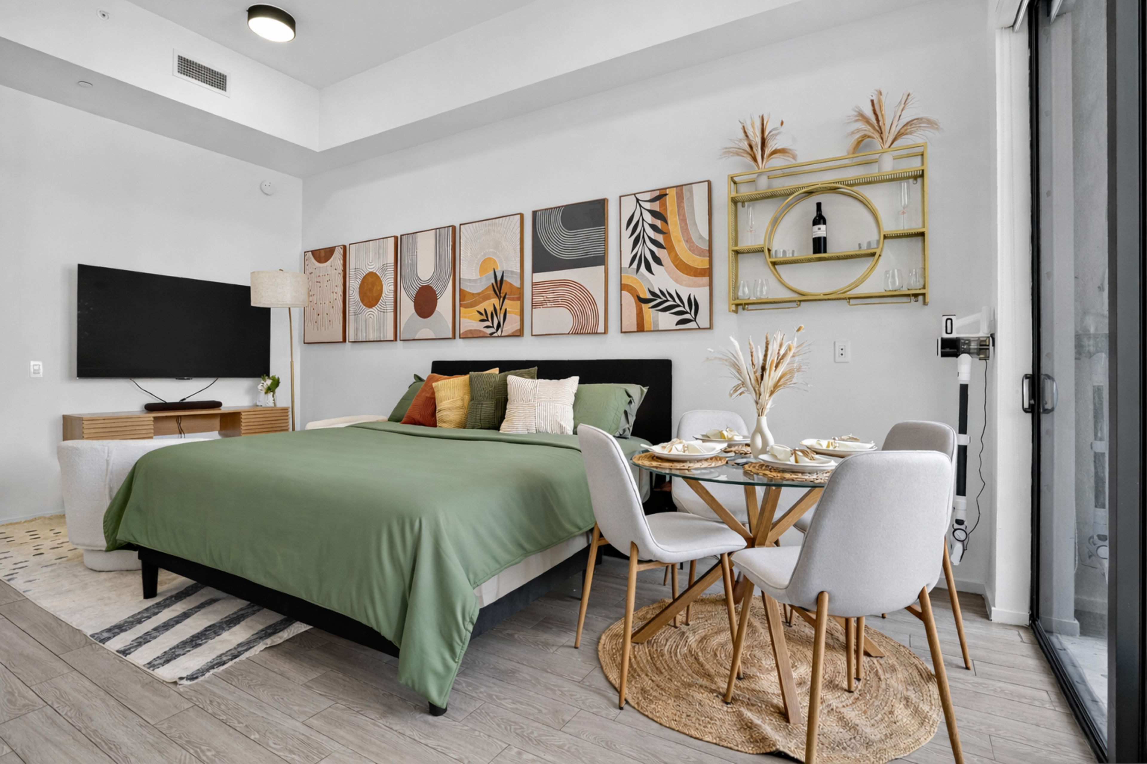 The image shows a modern studio bedroom featuring a bed with green bedding, a television mounted on the wall, and a small dining area with a round table and four chairs.
