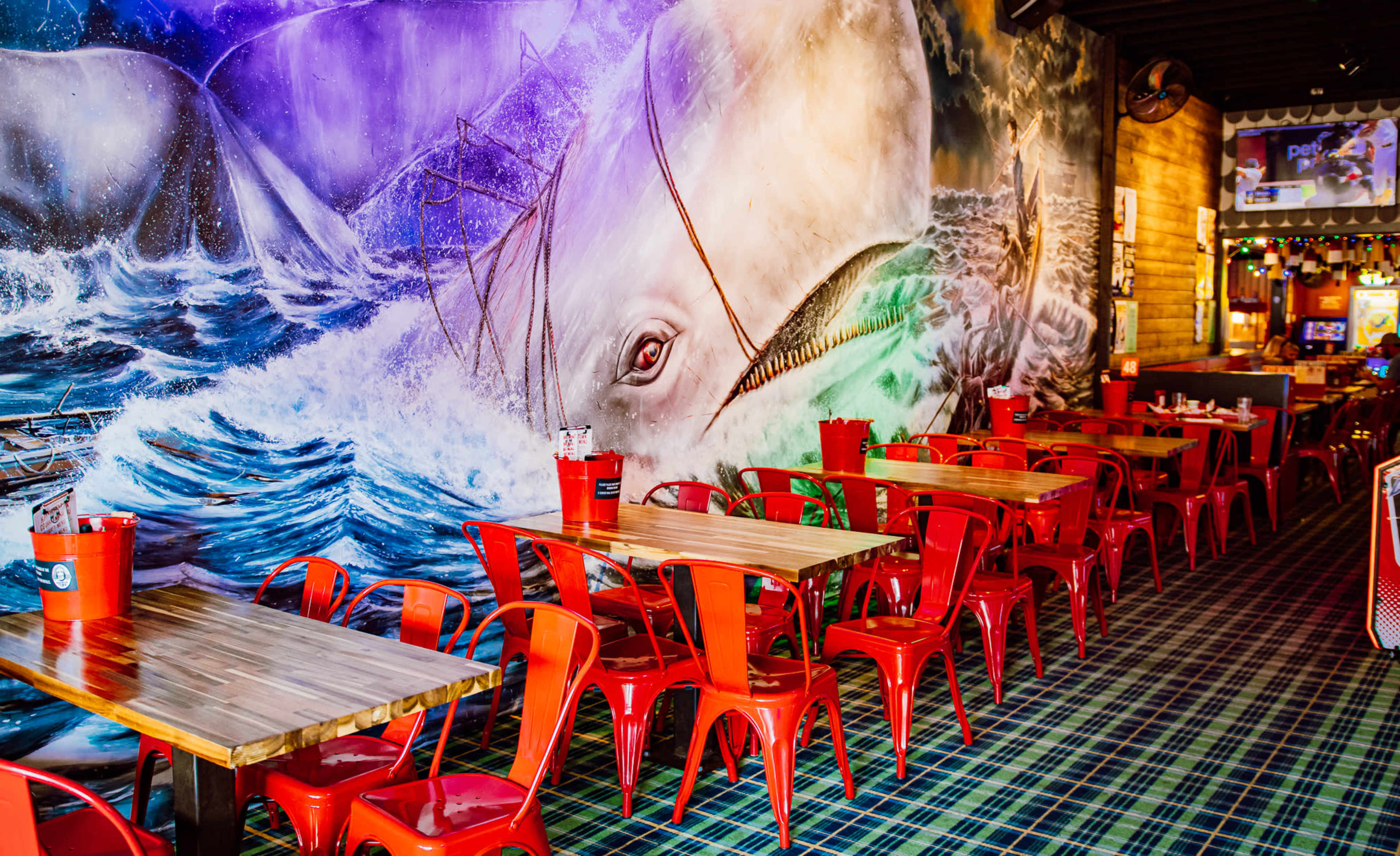 The image shows a restaurant interior with wooden tables and red metal chairs, featuring a large mural of a whale and turbulent ocean waves on the wall.