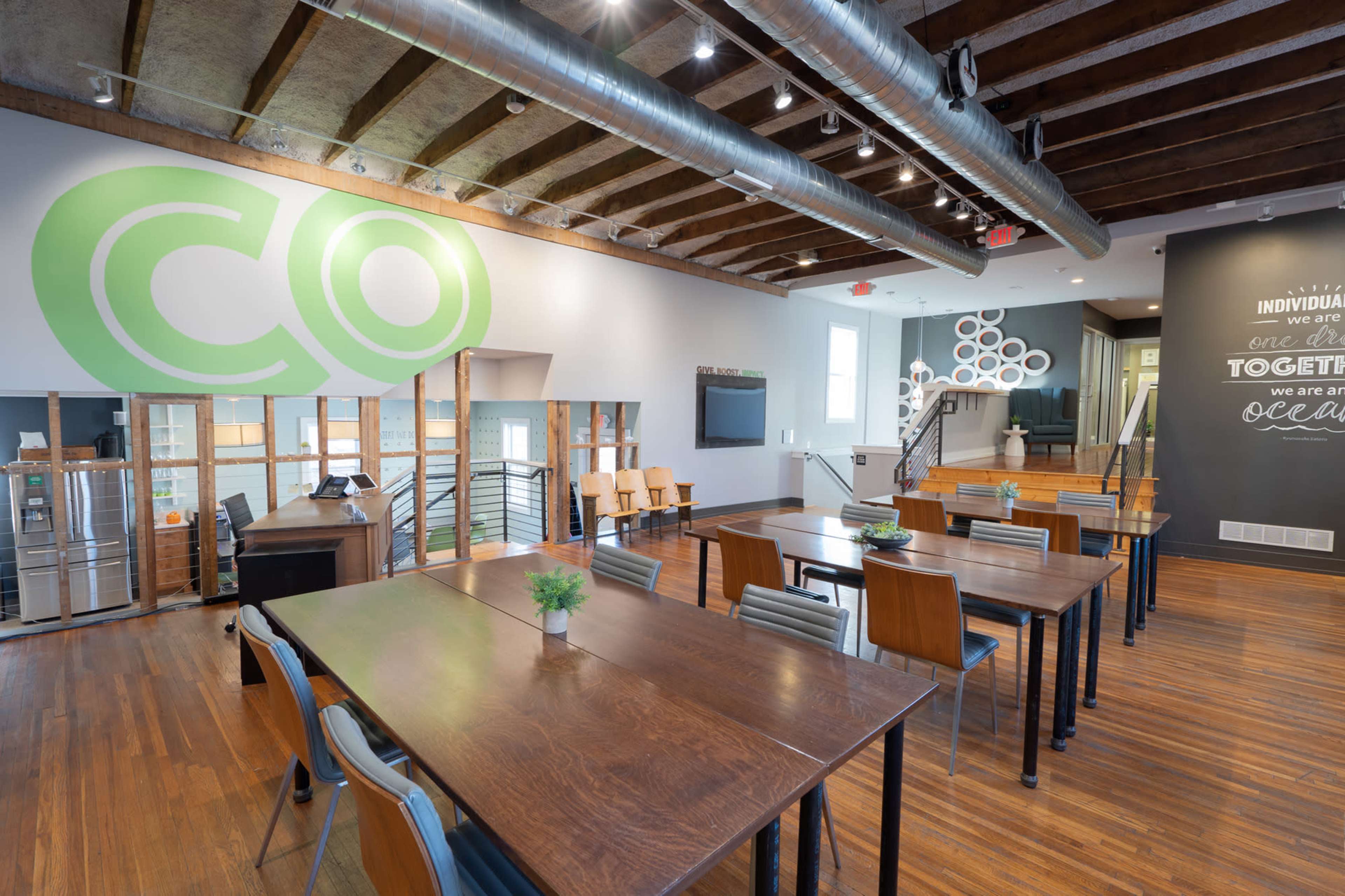 The image shows a spacious, well-lit room with wooden floors, long tables with chairs, and a large green logo on the wall, highlighting an open and modern design.